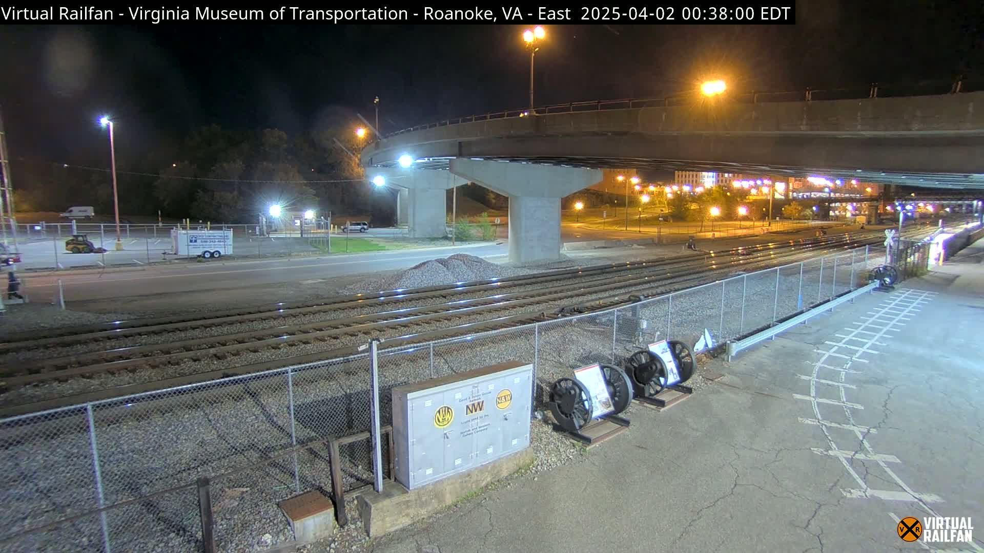 At night, under a highway overpass, railroad tracks extend toward a city with lights visible in the distance.