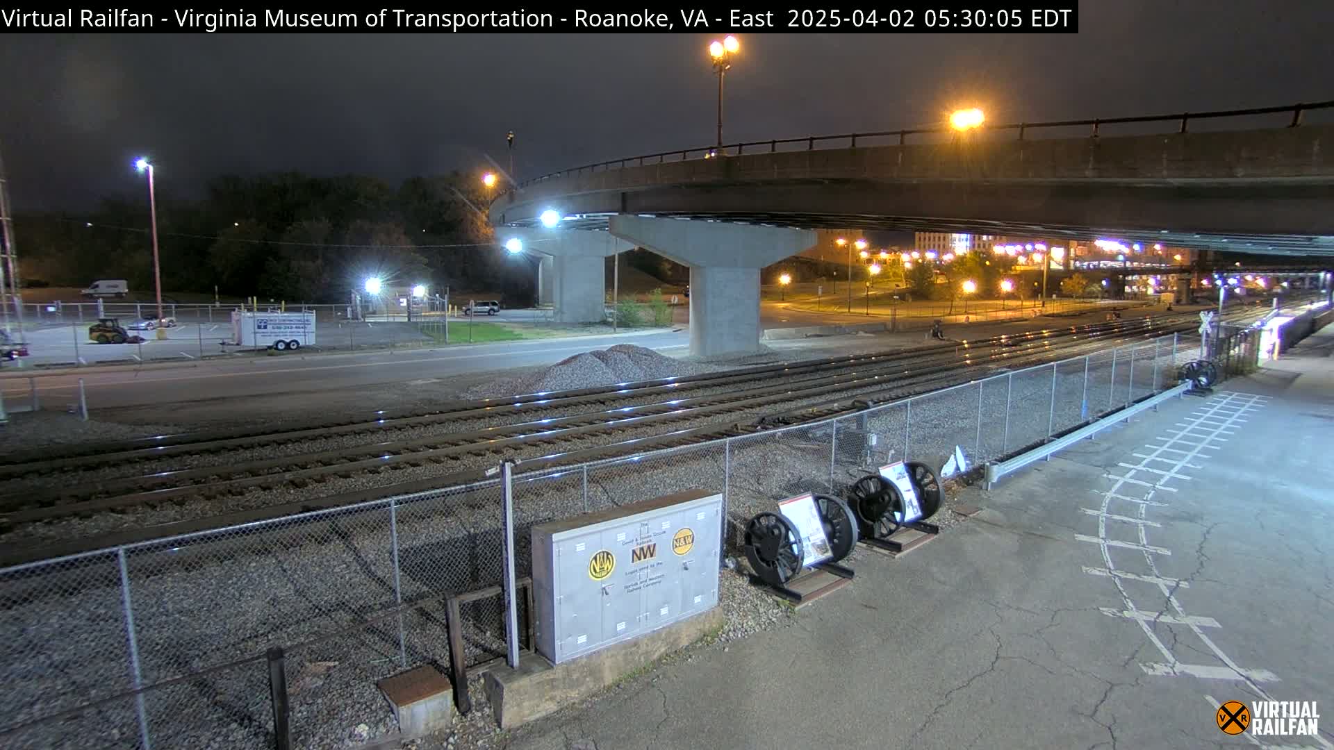 A nighttime view of railroad tracks and a roadway under a bridge, with a few vehicles visible in a parking lot.