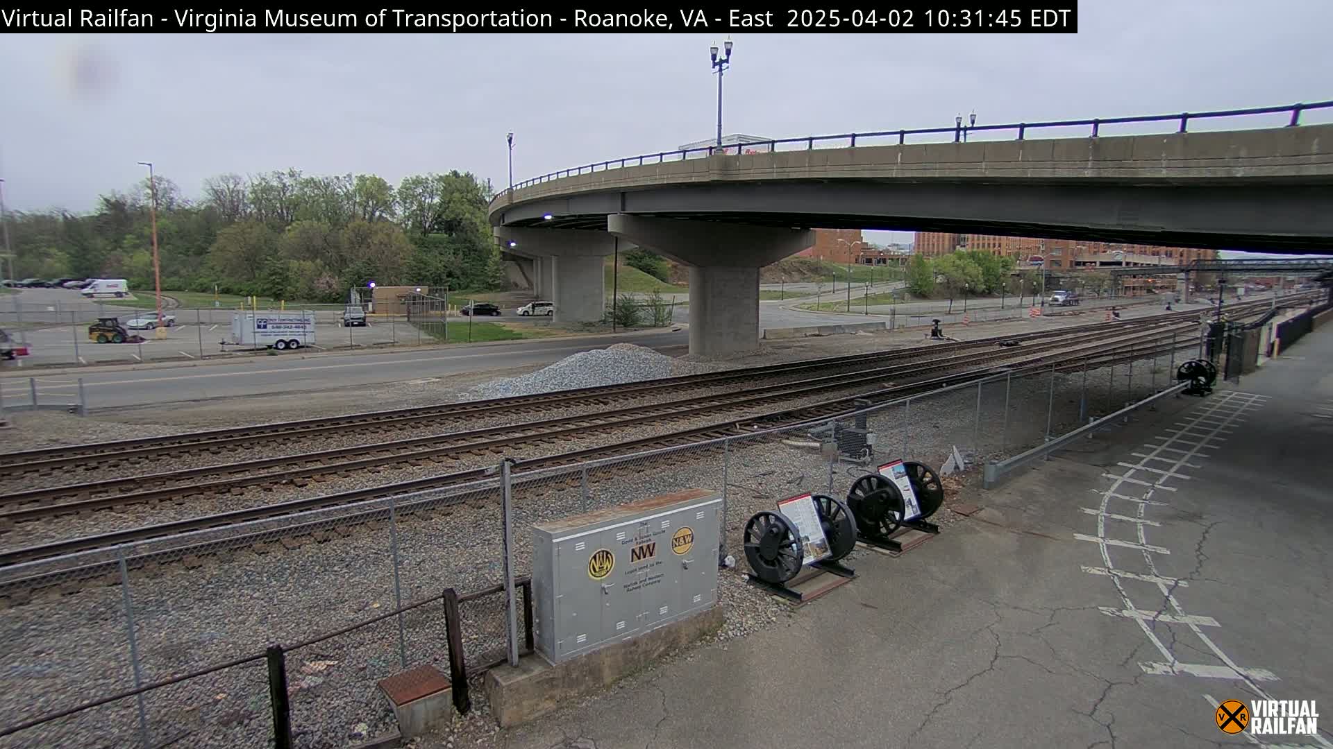 Under an overcast sky, railroad tracks run alongside a road that passes under a bridge, with a parking lot and some small buildings visible in the background.
