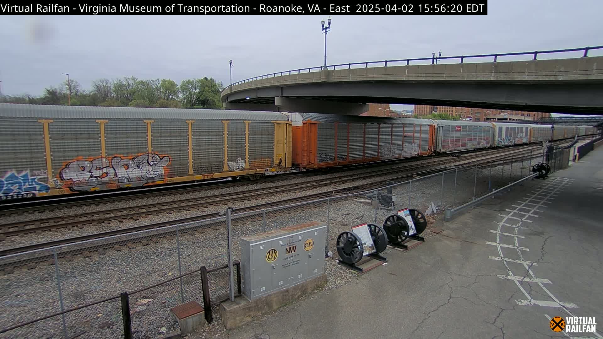 Under an overcast sky, a freight train with graffiti-covered cars passes under a bridge, alongside a fenced area with railway equipment and signage.