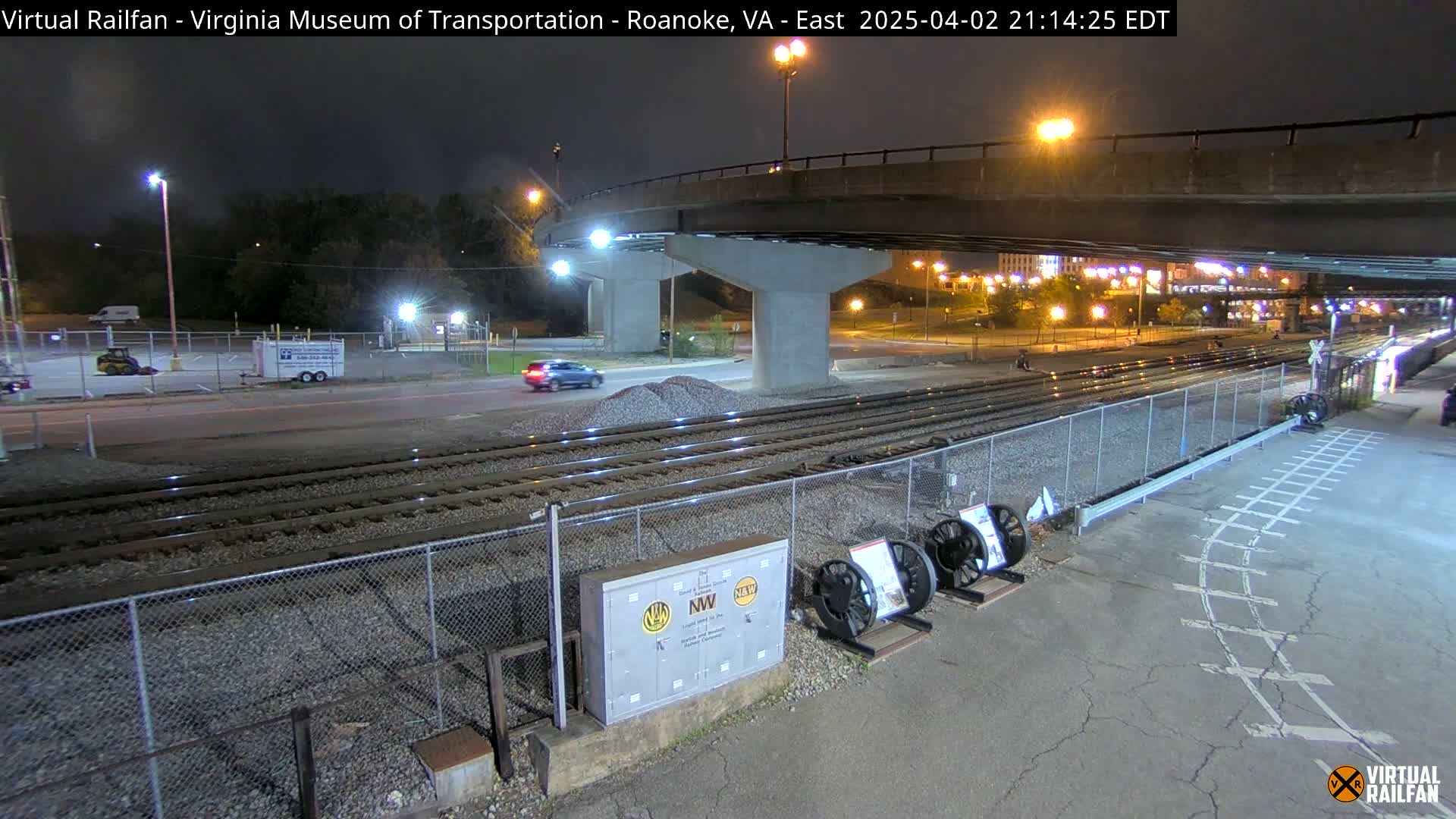 A nighttime view of railroad tracks and a roadway under a bridge, with a few vehicles visible in a parking lot and on the road.
