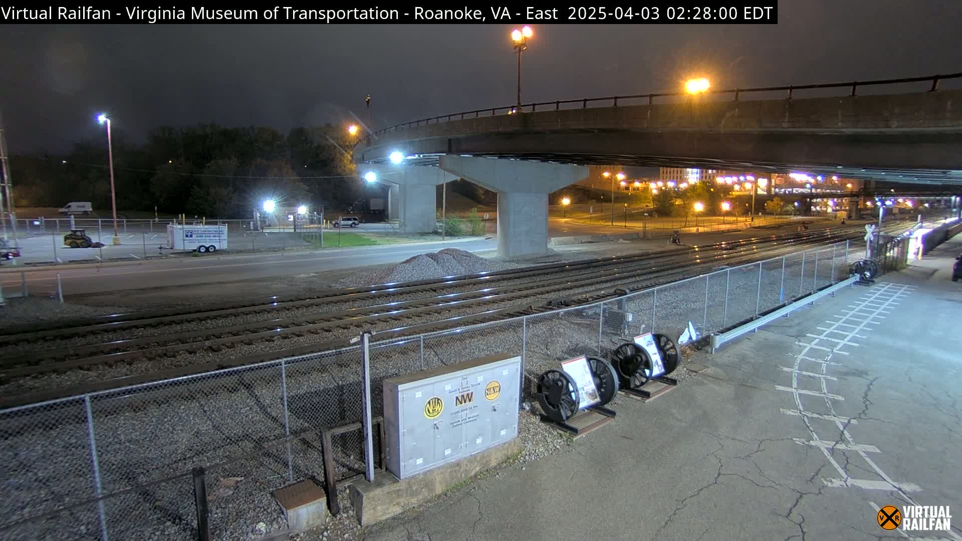 At night, under a bridge, railroad tracks run alongside a paved area next to a chain-link fence, with some pieces of railway equipment visible.
