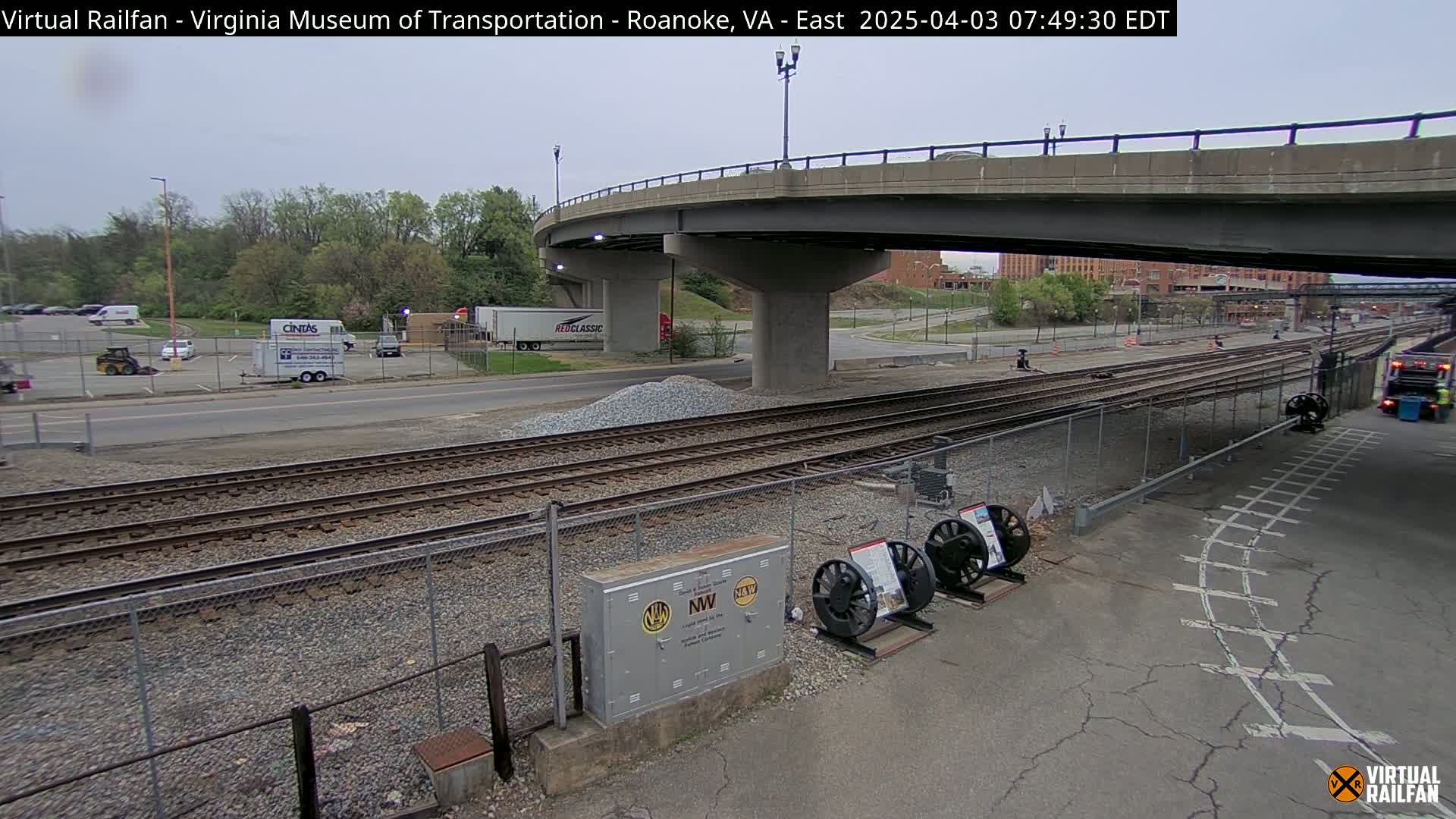 Under an overcast sky, railroad tracks run alongside a road, passing under a bridge; several vehicles are parked in a lot nearby.