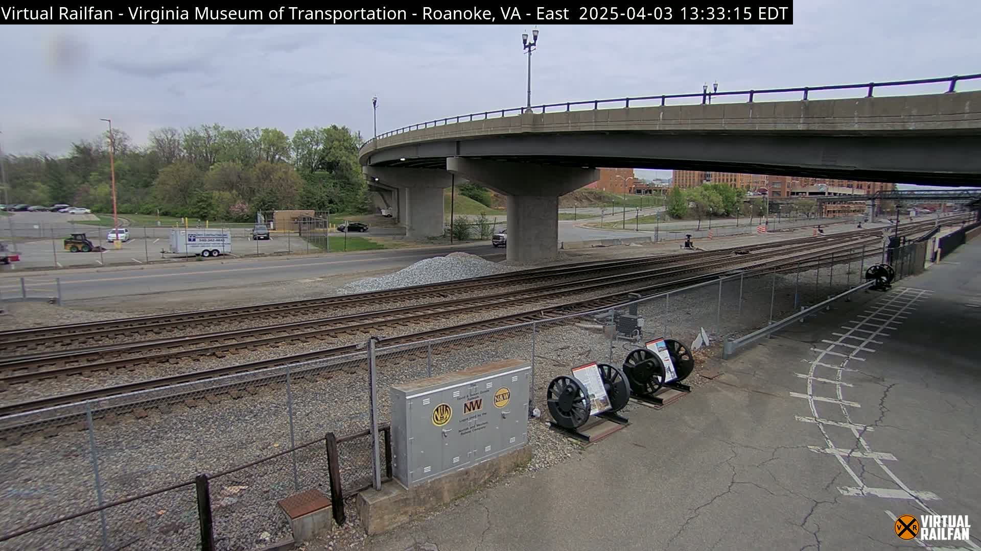 Here is a description of the image:

A mostly overcast day shows a view of railroad tracks next to a road, under a bridge, with a parking lot and some trees in the background.