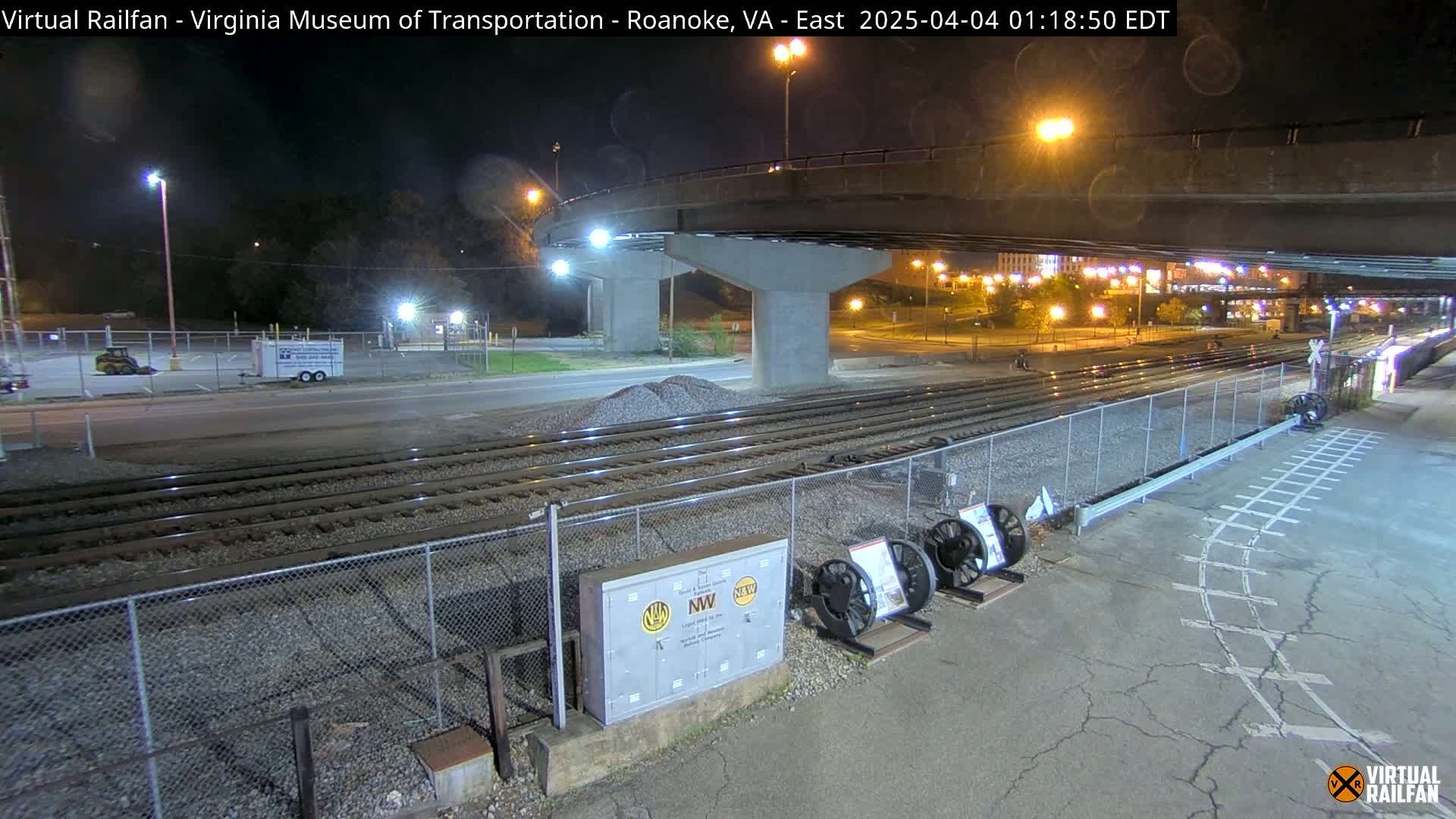 At night, under a bridge, railroad tracks extend toward a roadway, with some small vehicles visible in a parking lot.
