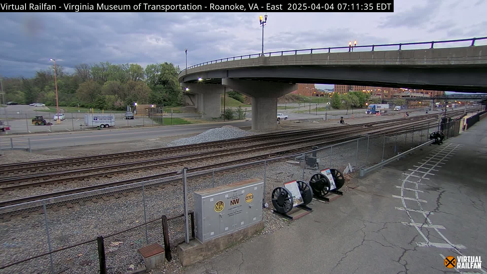 Under an overcast sky, railroad tracks extend beneath a bridge, with a parking lot and some trees visible in the background.