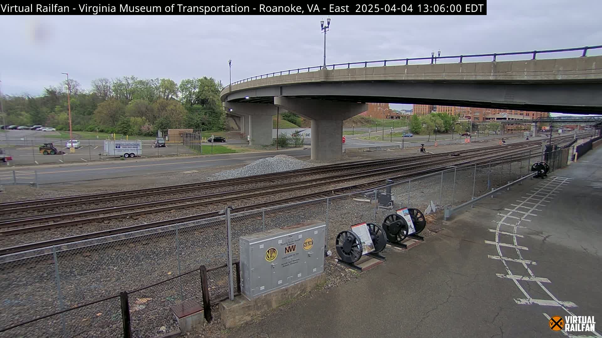 A view of railroad tracks under a bridge on an overcast day, with a parking lot and some vehicles visible in the background.