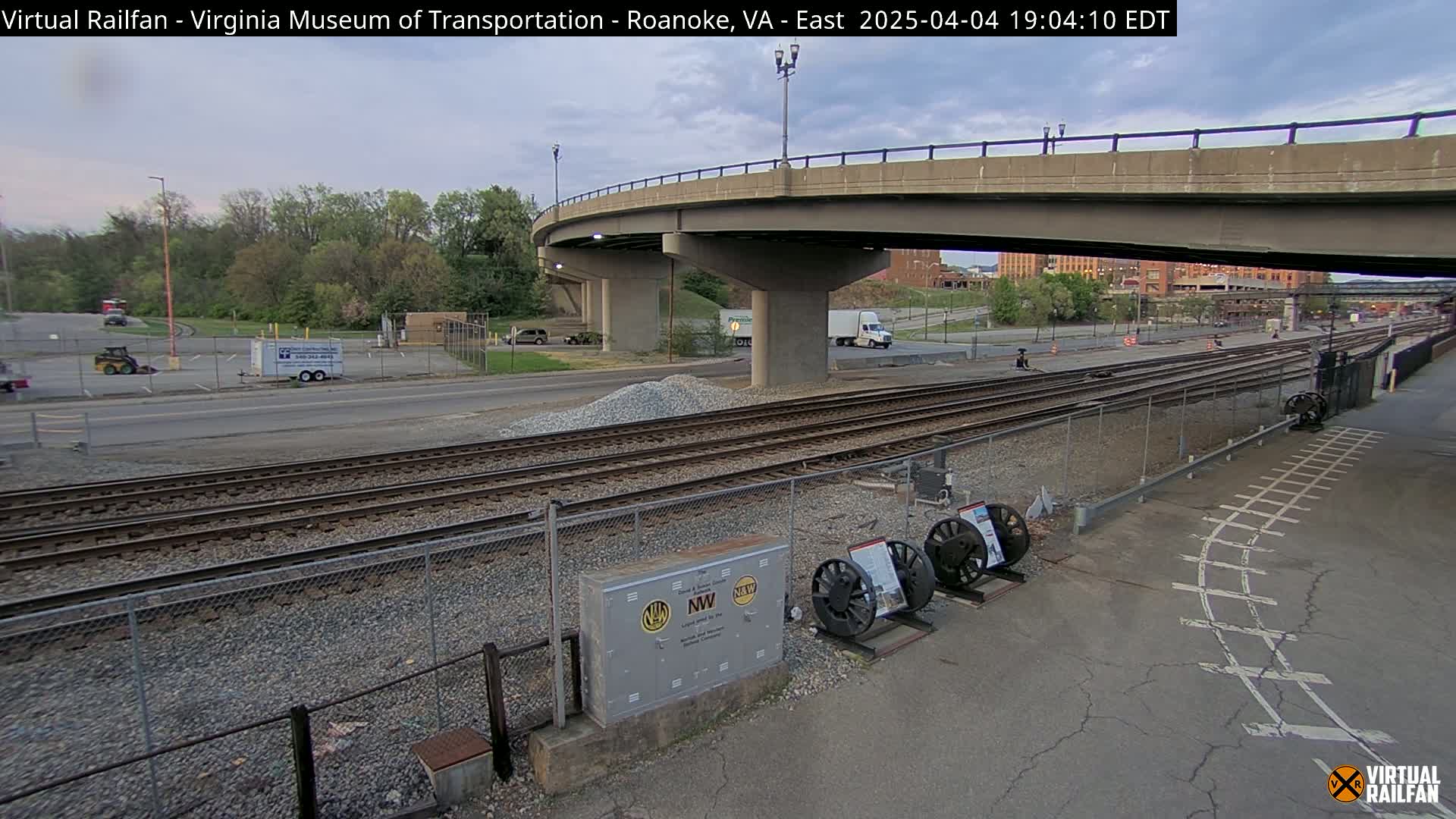A cloudy outdoor scene shows railroad tracks next to a road, under a bridge, with some vehicles and construction-related materials visible.