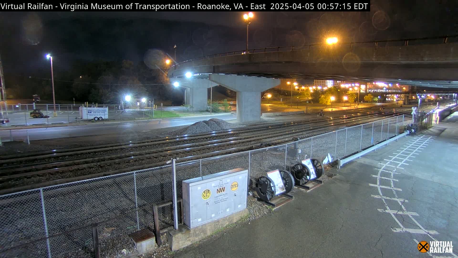 A nighttime view of railroad tracks beneath a highway overpass, with a chain-link fence separating the tracks from a paved area containing some equipment.