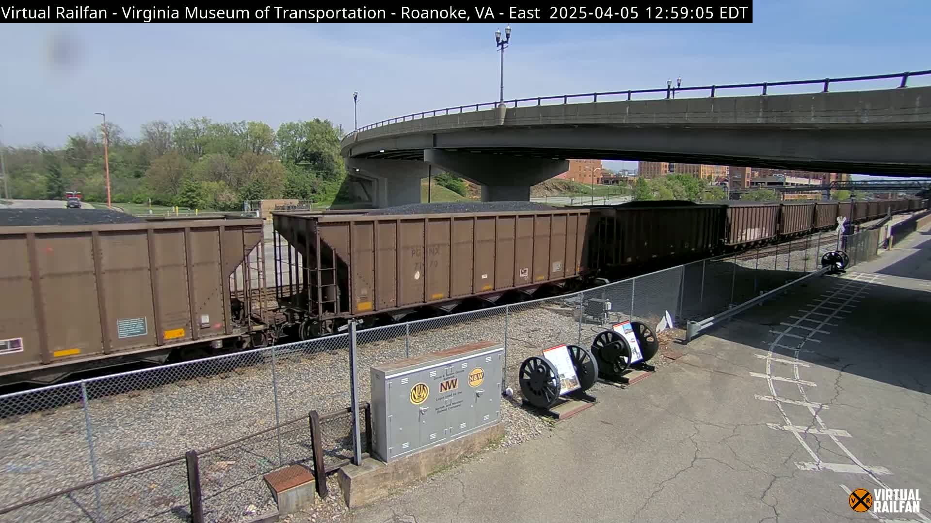 A train carrying coal sits under a bridge on a sunny day.