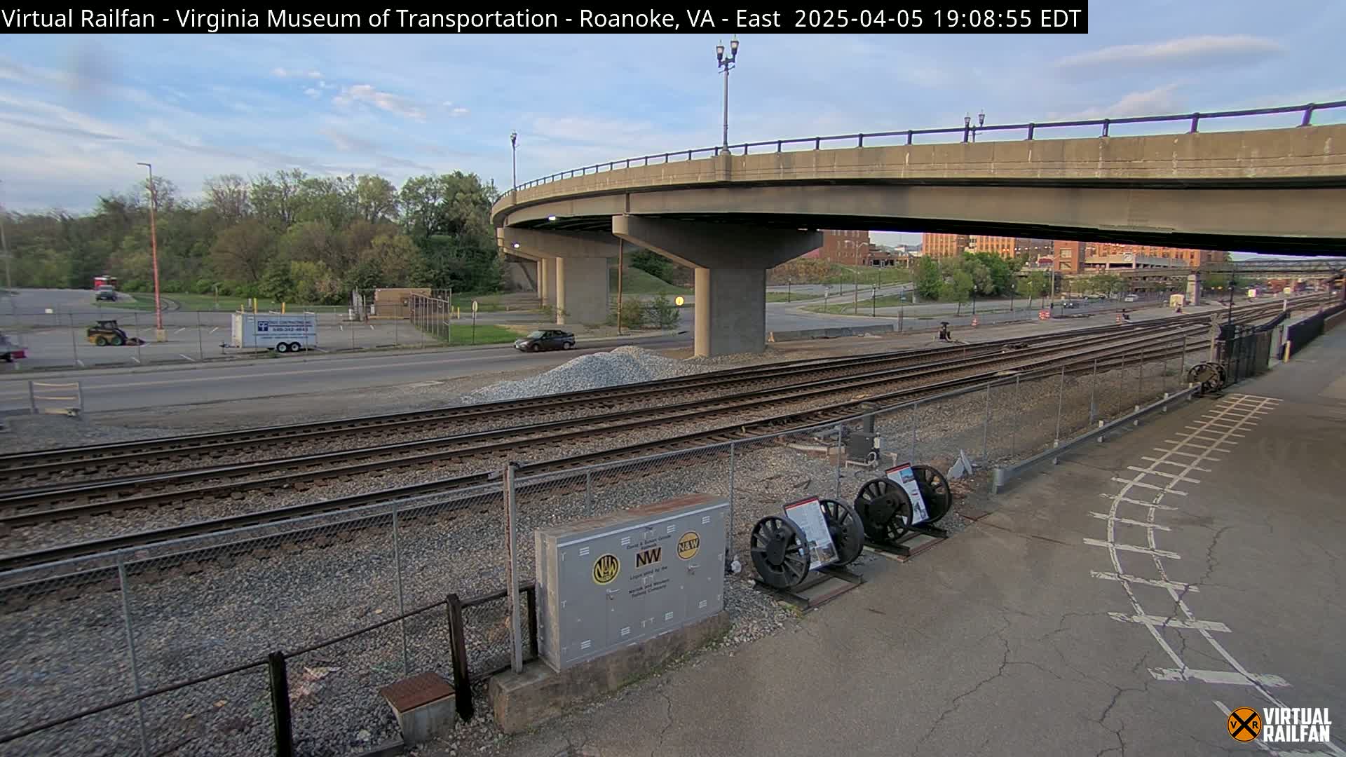 Under a partly cloudy evening sky, railroad tracks run alongside a road beneath a curved overpass, with some vehicles visible in the distance and a few pieces of railway equipment nearby.