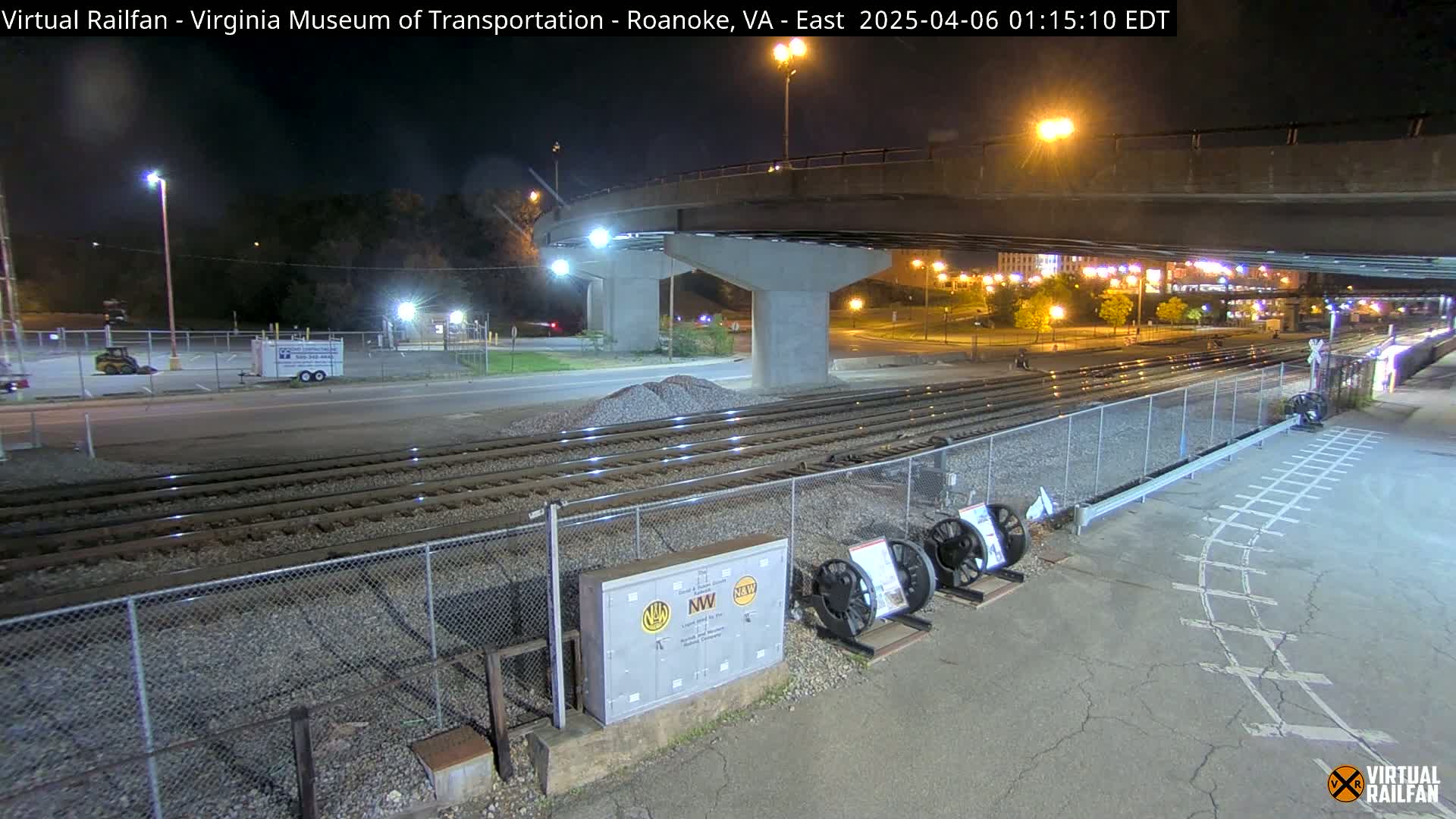 A nighttime view of railroad tracks next to a road, under a highway overpass, with some parked vehicles and equipment visible in the distance.