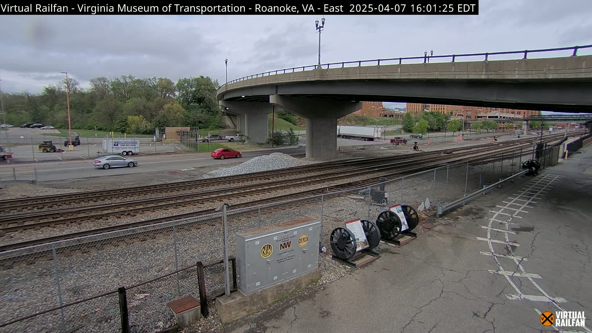 A mostly cloudy day shows a view of railroad tracks under a bridge, with several cars and trucks visible on a road nearby.
