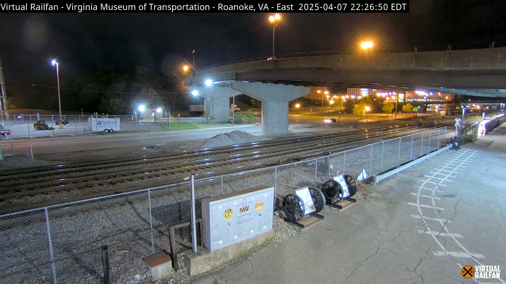 Under a nighttime sky, railroad tracks extend beneath a bridge, with a roadway visible beyond a chain-link fence.