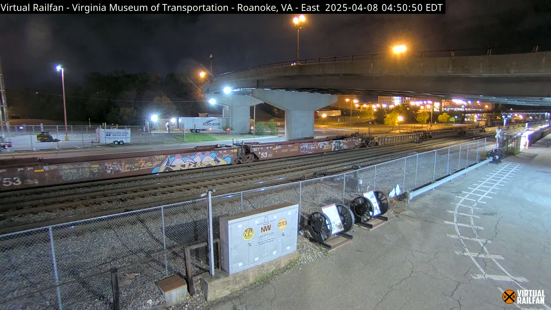 At night, under a highway overpass, a freight train with graffiti-covered cars sits on railroad tracks near a fenced-in area with some equipment.