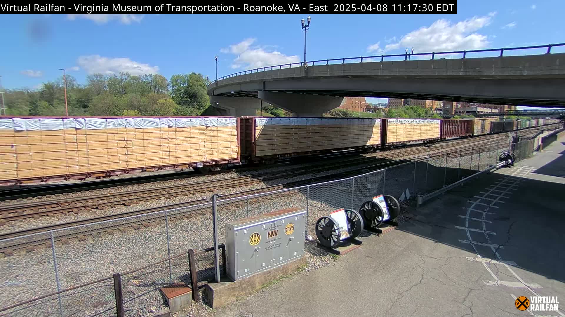 A long freight train carrying lumber passes under a bridge on a partly sunny day.
