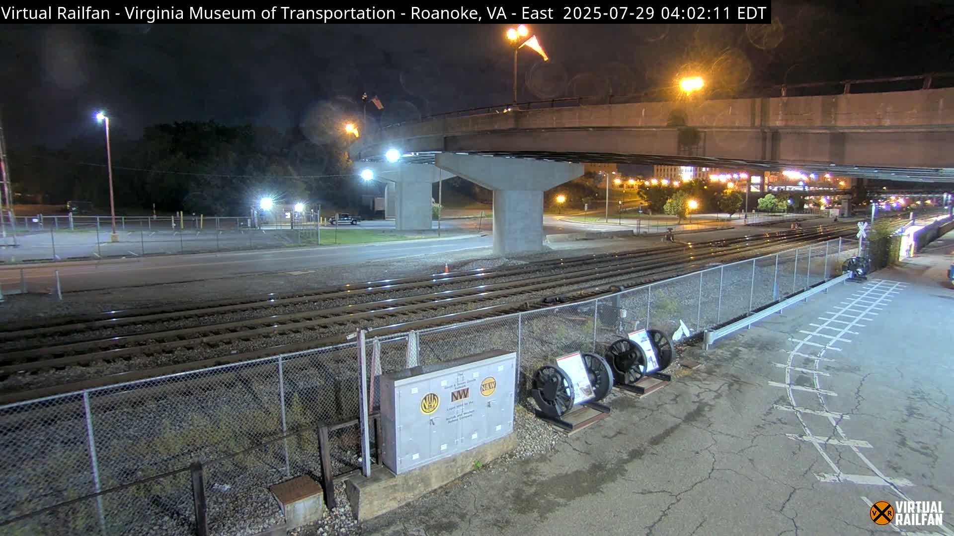 At night, a roadway and railroad tracks intersect beneath a highway overpass, with several train wheels and a metal box visible near the tracks.