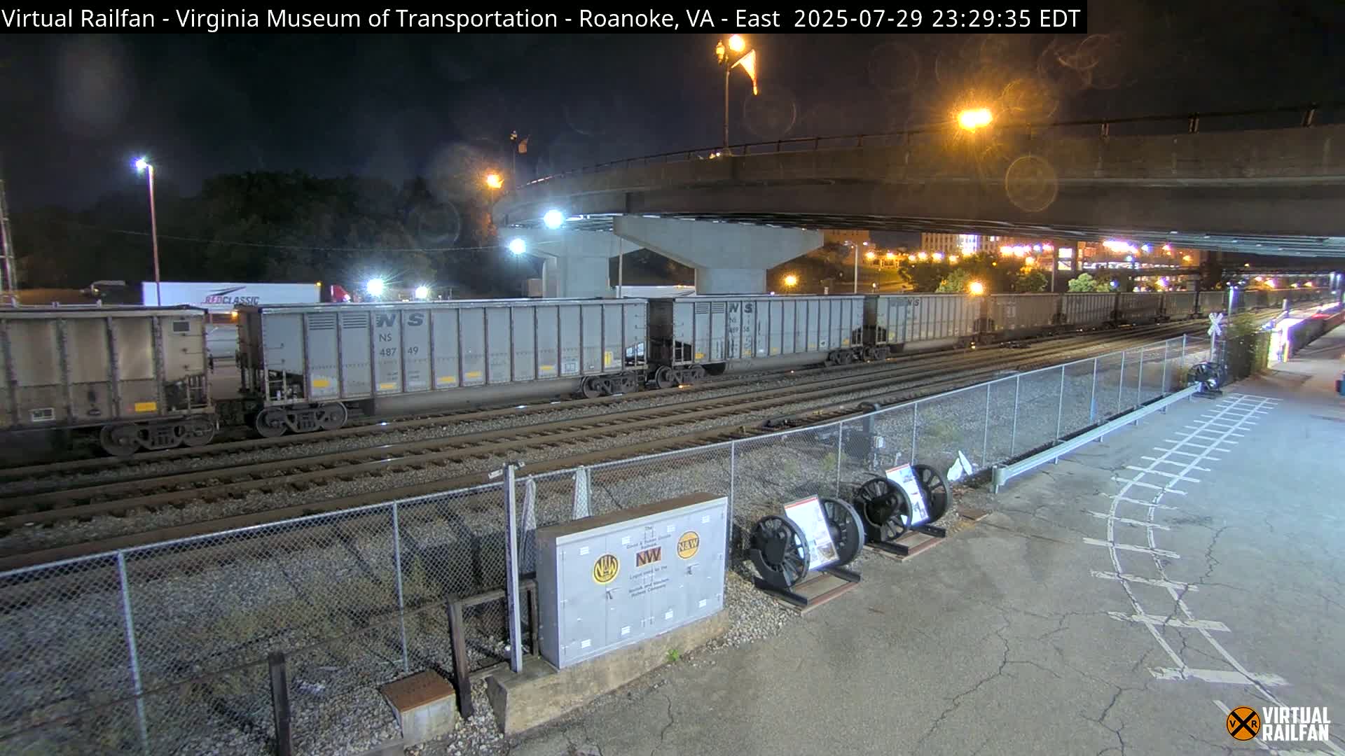At night, a roadway and railroad tracks intersect beneath a highway overpass, with several train wheels and a metal box visible near the tracks.