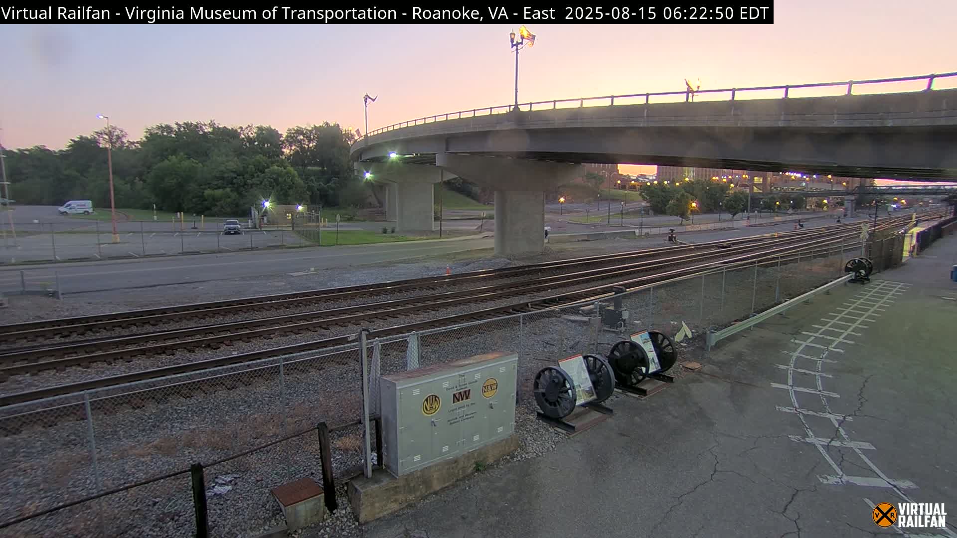 At night, a roadway and railroad tracks intersect beneath a highway overpass, with several train wheels and a metal box visible near the tracks.