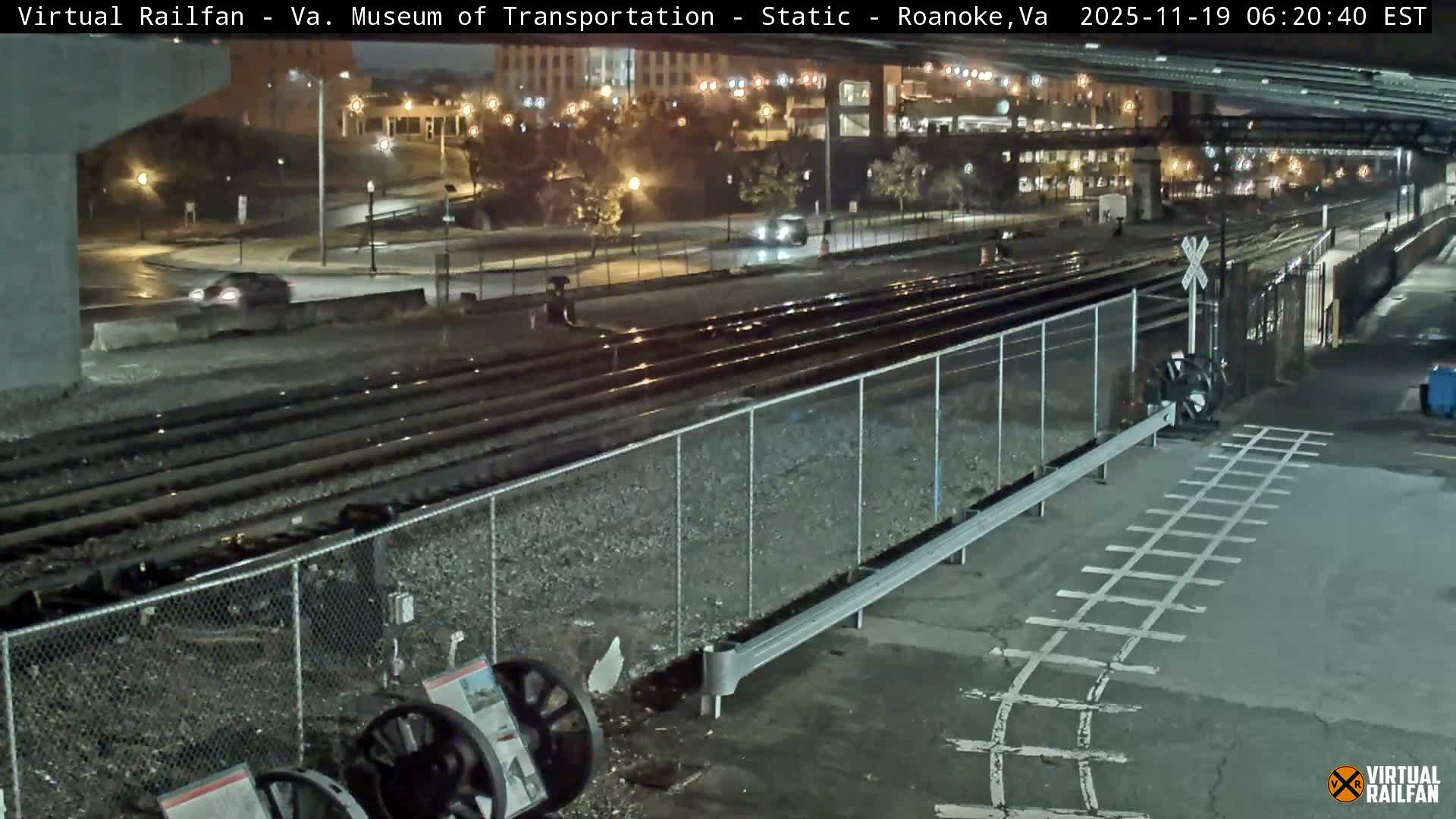 At night, a roadway and railroad tracks intersect beneath a highway overpass, with several train wheels and a metal box visible near the tracks.