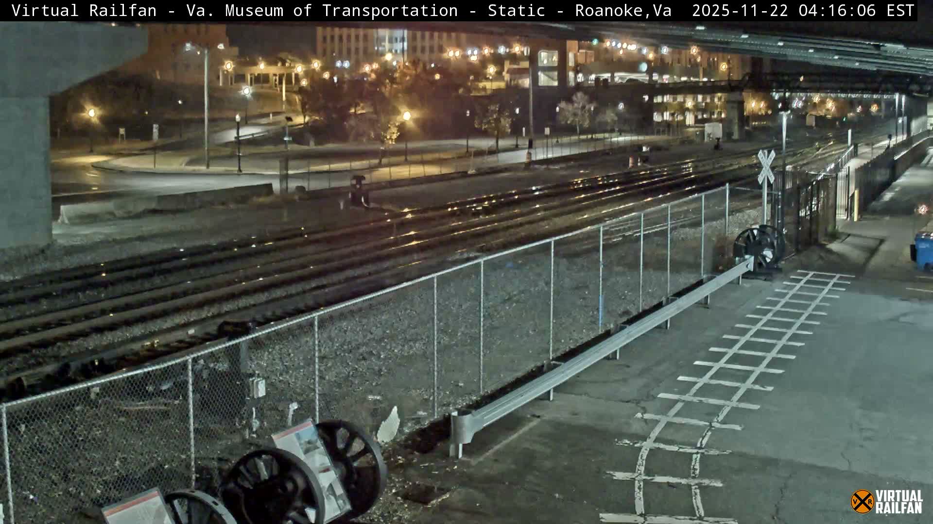 Multiple railroad tracks run through an urban area at night, separated by a chain-link fence from a paved pedestrian area with a painted track crossing, all appearing damp from recent moisture.