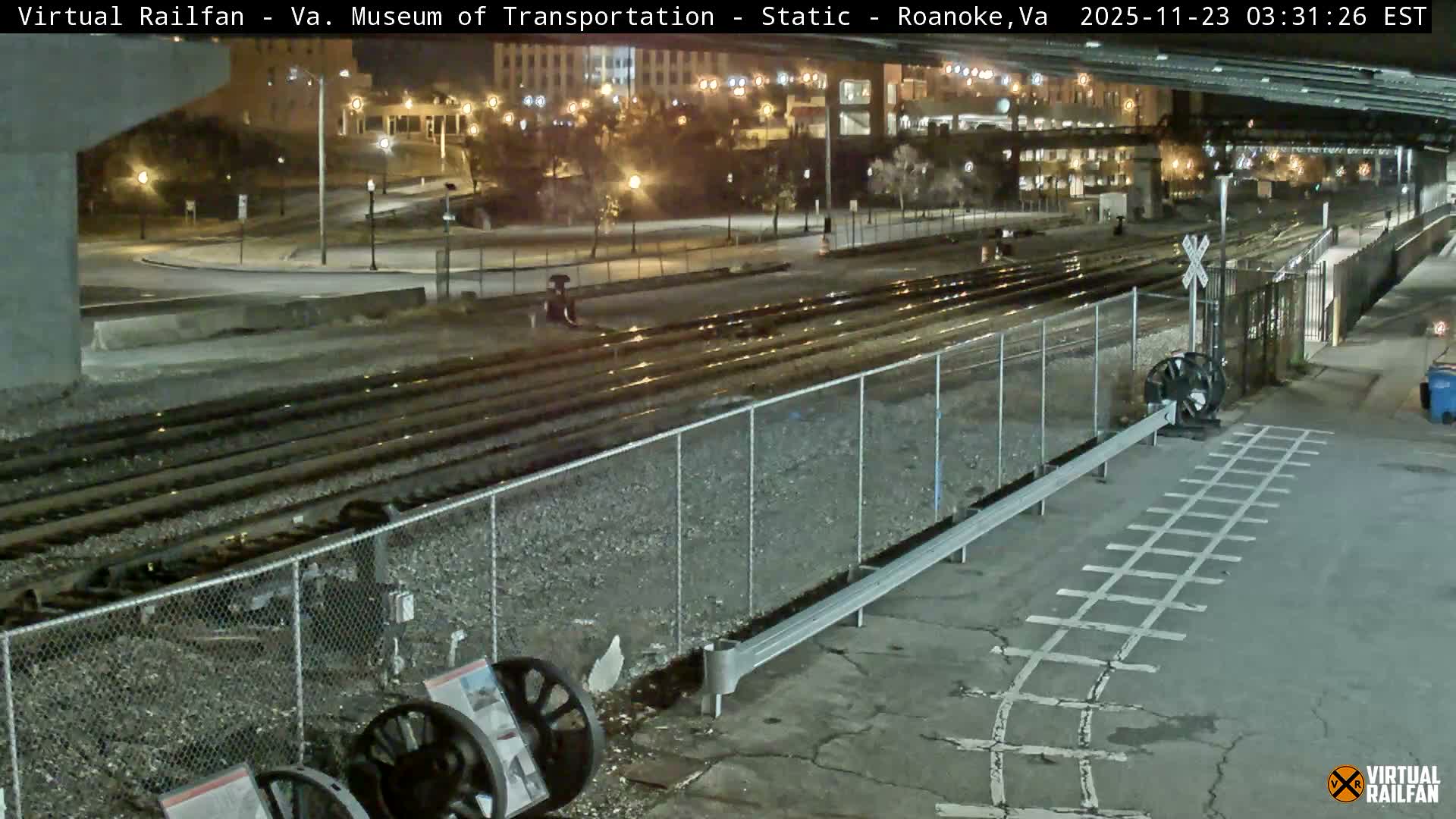 Multiple railroad tracks and a pedestrian crossing are visible at night, with wet ground reflecting city lights from a nearby urban area and an overpass.