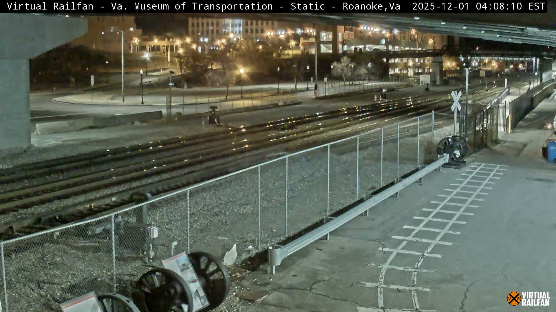 An outdoor nighttime view features multiple train tracks, a railroad crossing with a gate and painted markings, and large exhibit wheels in the foreground, all illuminated by numerous streetlights and distant building lights under clear weather.