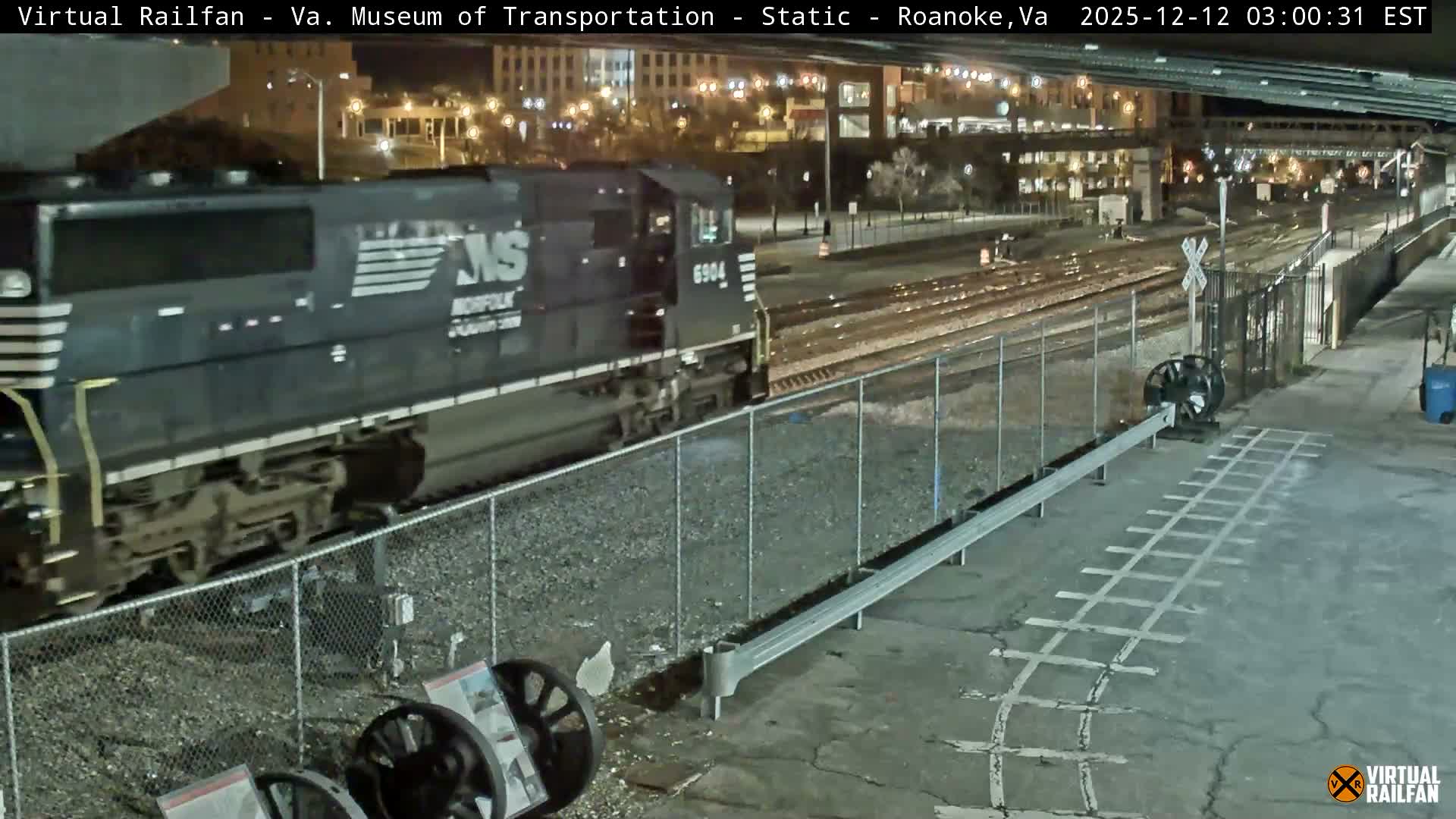A dark blue Norfolk Southern train locomotive, numbered 6904, moves past a station platform and multiple tracks at night, with city lights and an overpass visible in the background under clear weather conditions.