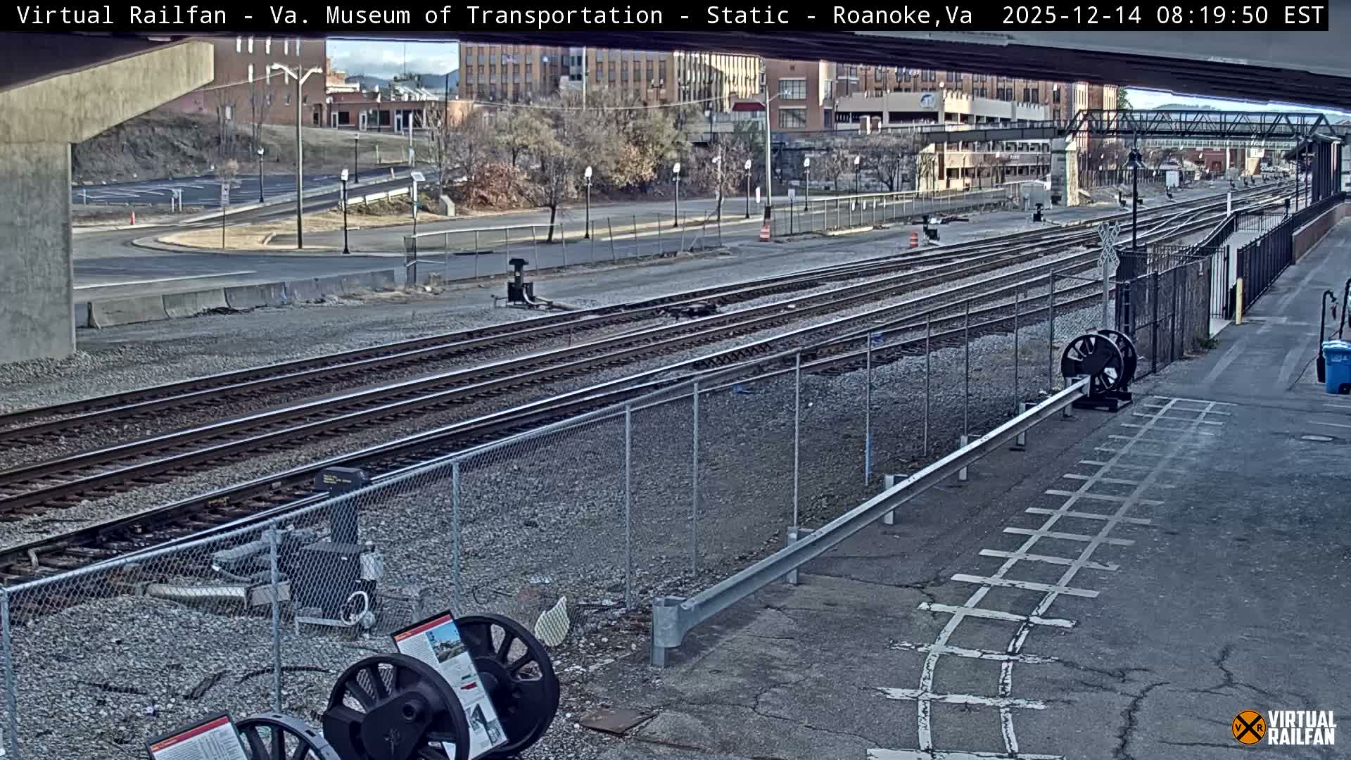 A dark blue Norfolk Southern train locomotive, numbered 6904, moves past a station platform and multiple tracks at night, with city lights and an overpass visible in the background under clear weather conditions.