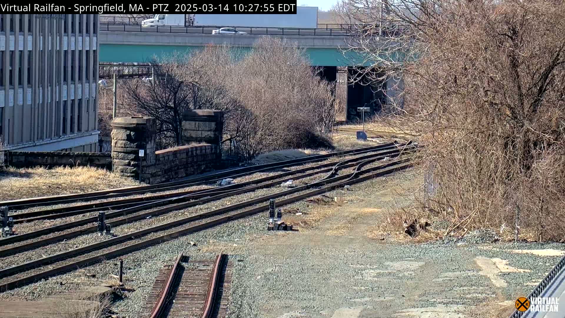 A sunny view of a railroad switching yard with several tracks, a stone wall, and a building in the background, under a highway overpass.