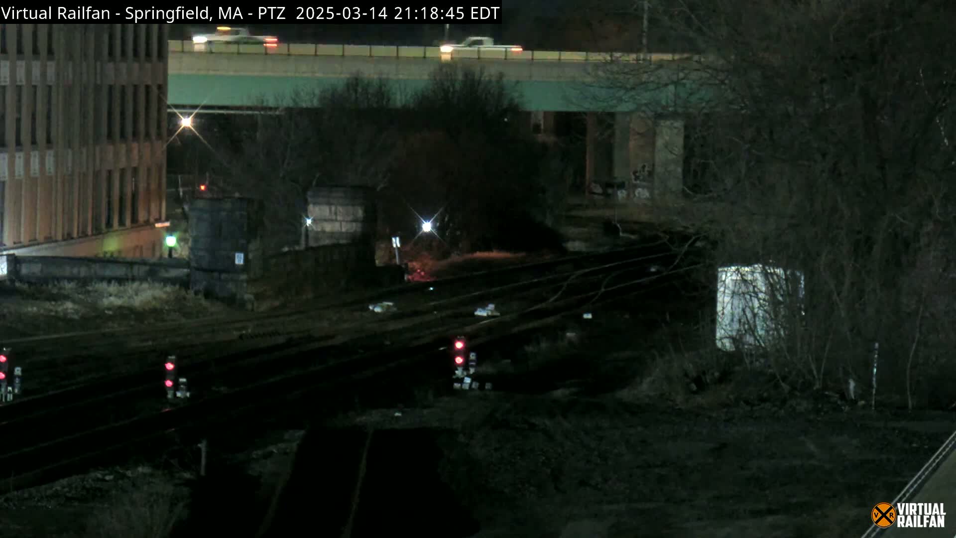 A nighttime view of railroad tracks under a highway overpass, near a multi-story building, with bare trees and dim lighting.