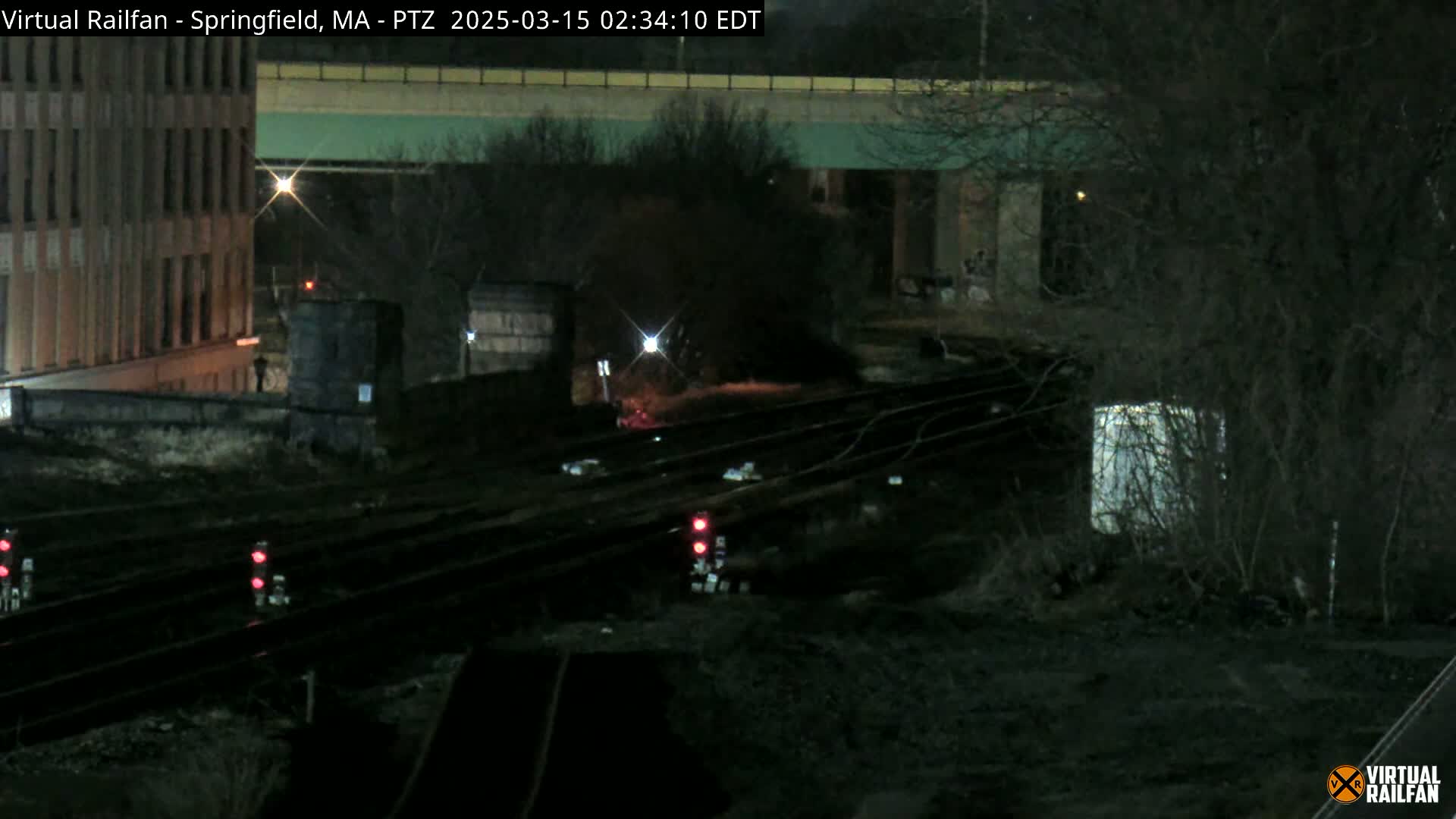 A nighttime view of railroad tracks under a highway overpass, with a building visible in the background and sparse vegetation surrounding the tracks.