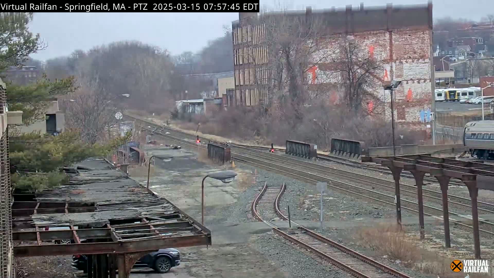 A view of railroad tracks and switching areas on an overcast day, with a partly dilapidated structure in the foreground,  bare trees, and brick buildings in the background.