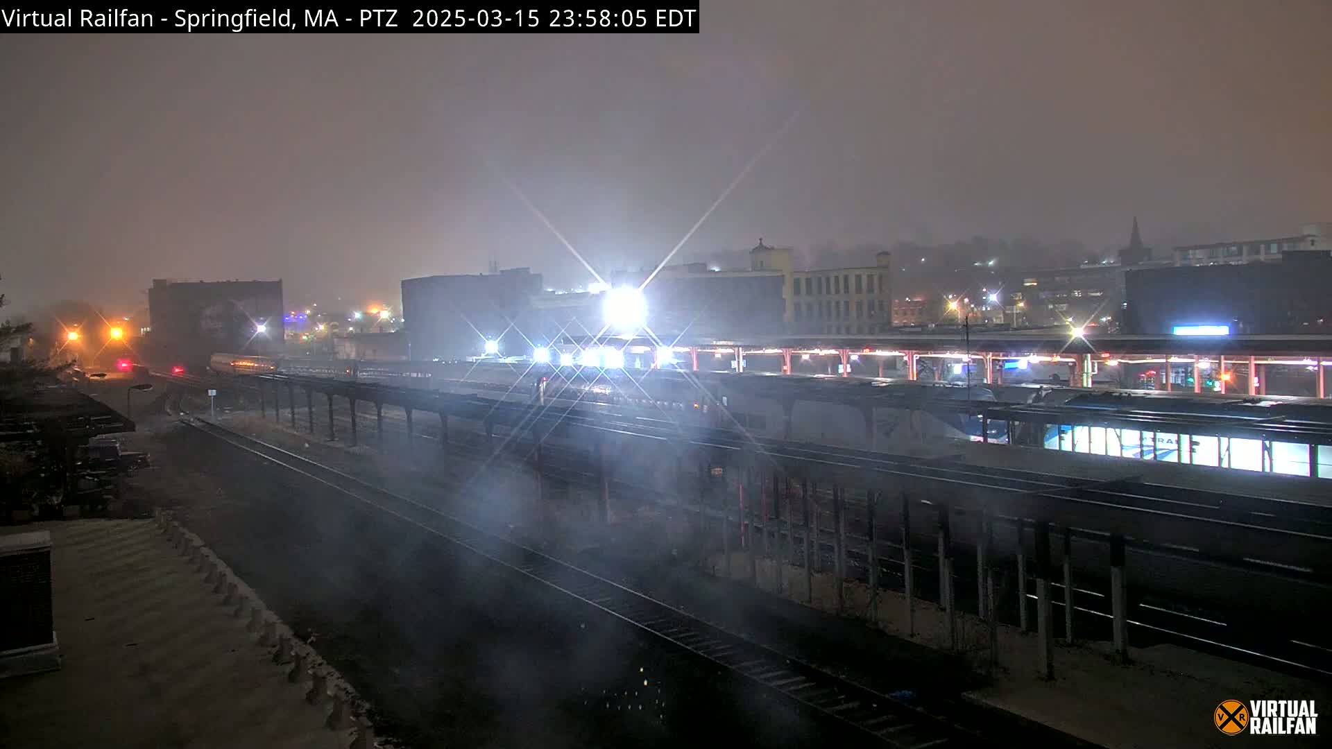 A nighttime view of a train station in snowy conditions, showing trains at the platforms and tracks extending into the foggy distance.