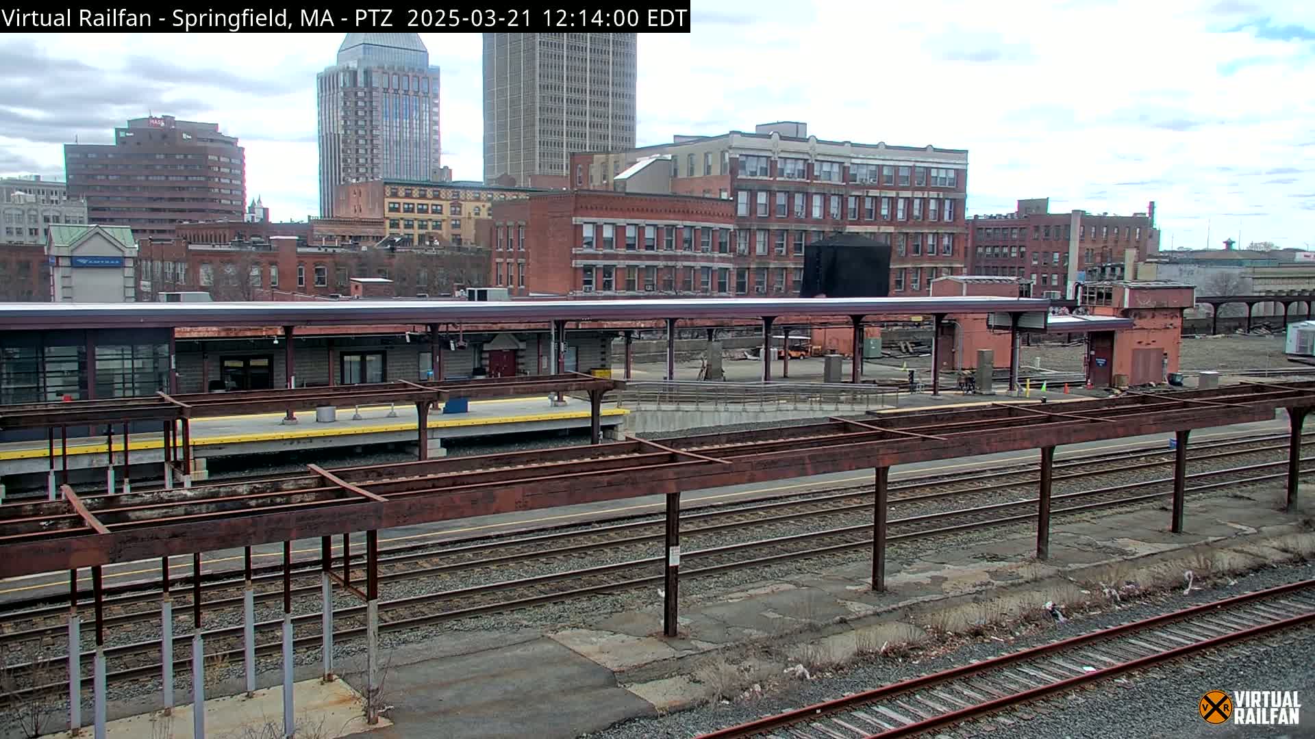 A train station platform with a covered waiting area and multiple train tracks sits before a city skyline under a partly cloudy sky.