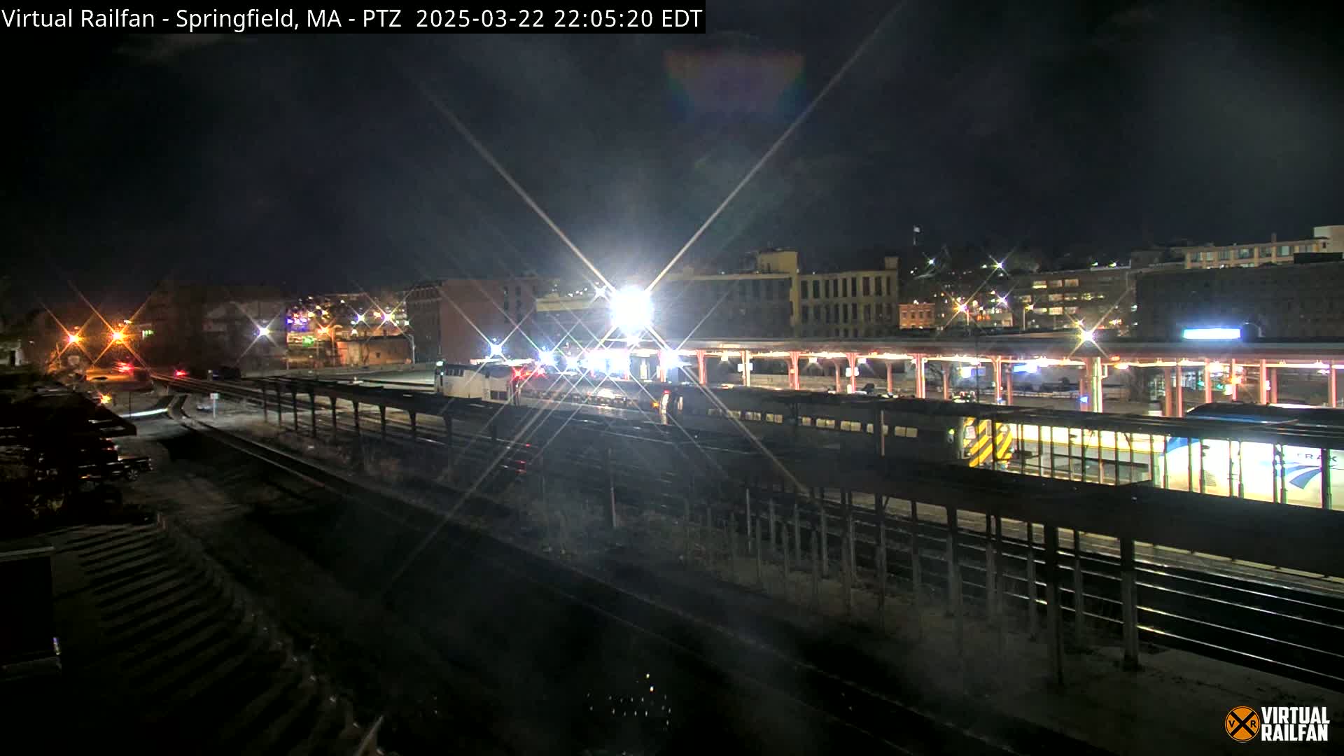 A nighttime view of a train station with a train illuminated by bright lights, under a dark sky.