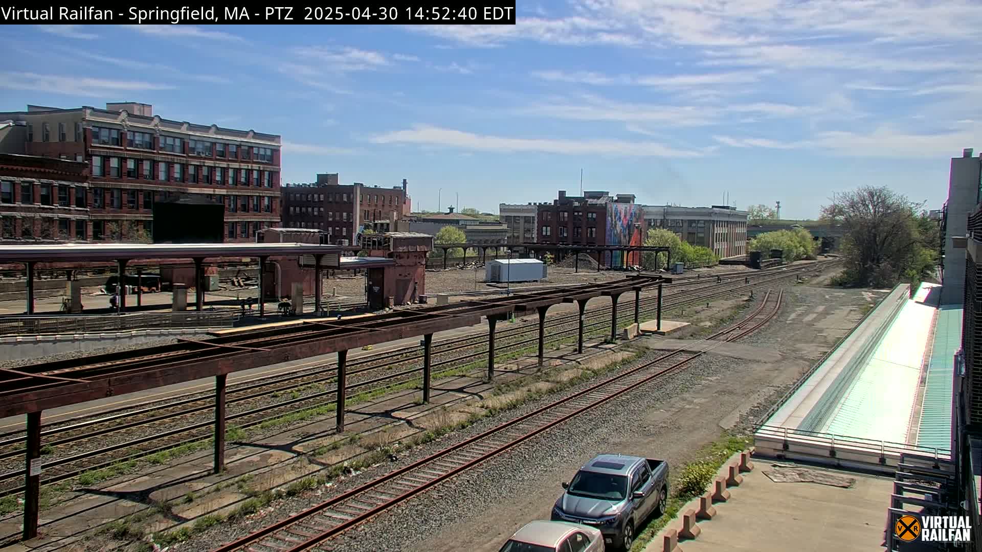 A sunny day reveals a railway yard with multiple train tracks, brick buildings, and a partially covered platform, all under a mostly clear sky.