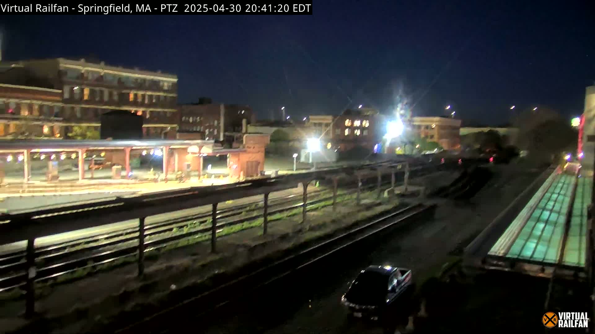 A nighttime view of a train station with several buildings in the background under a clear, dark sky.