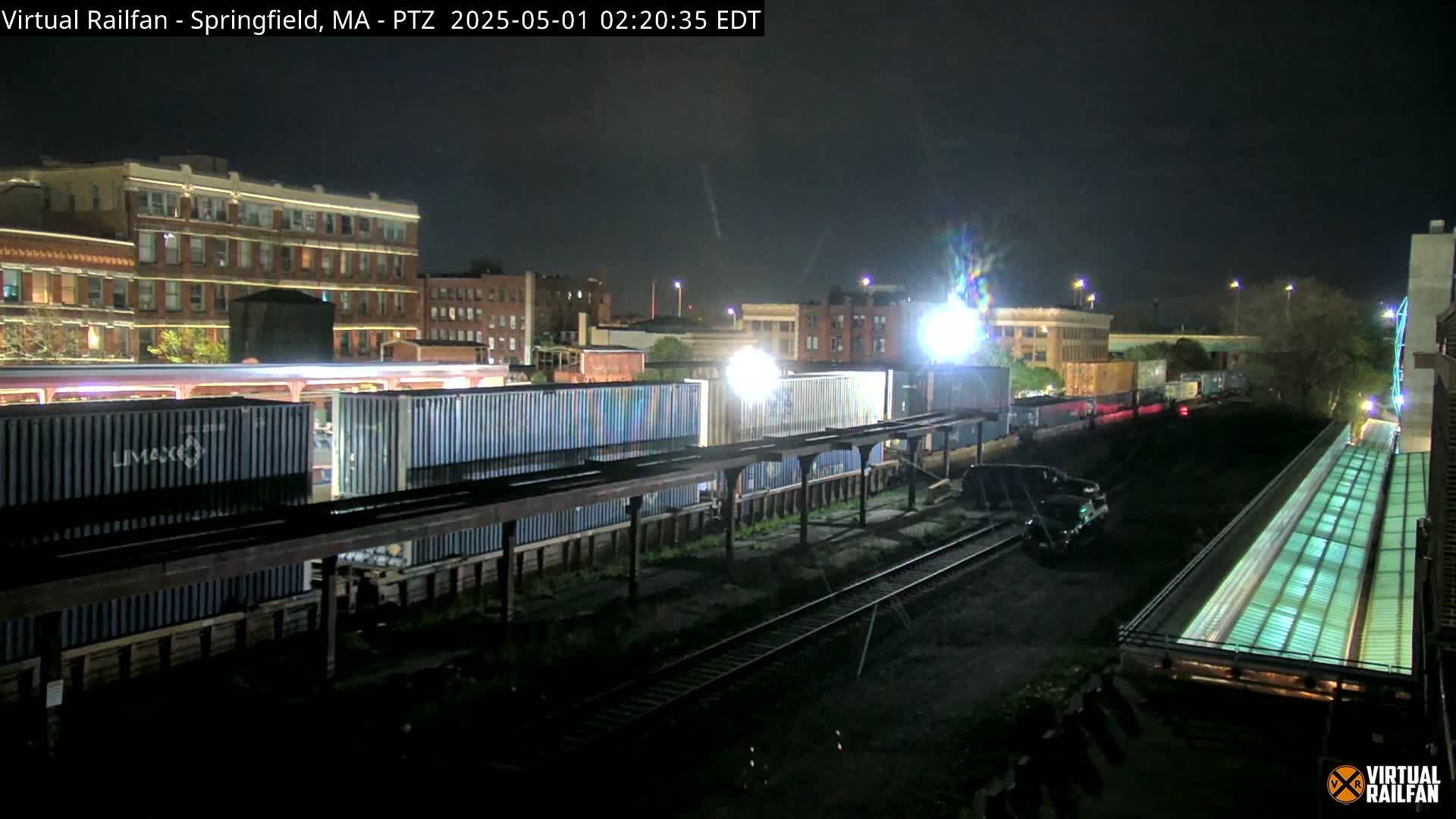 A nighttime view of a train yard with several shipping containers, railway tracks, and buildings illuminated by artificial light.