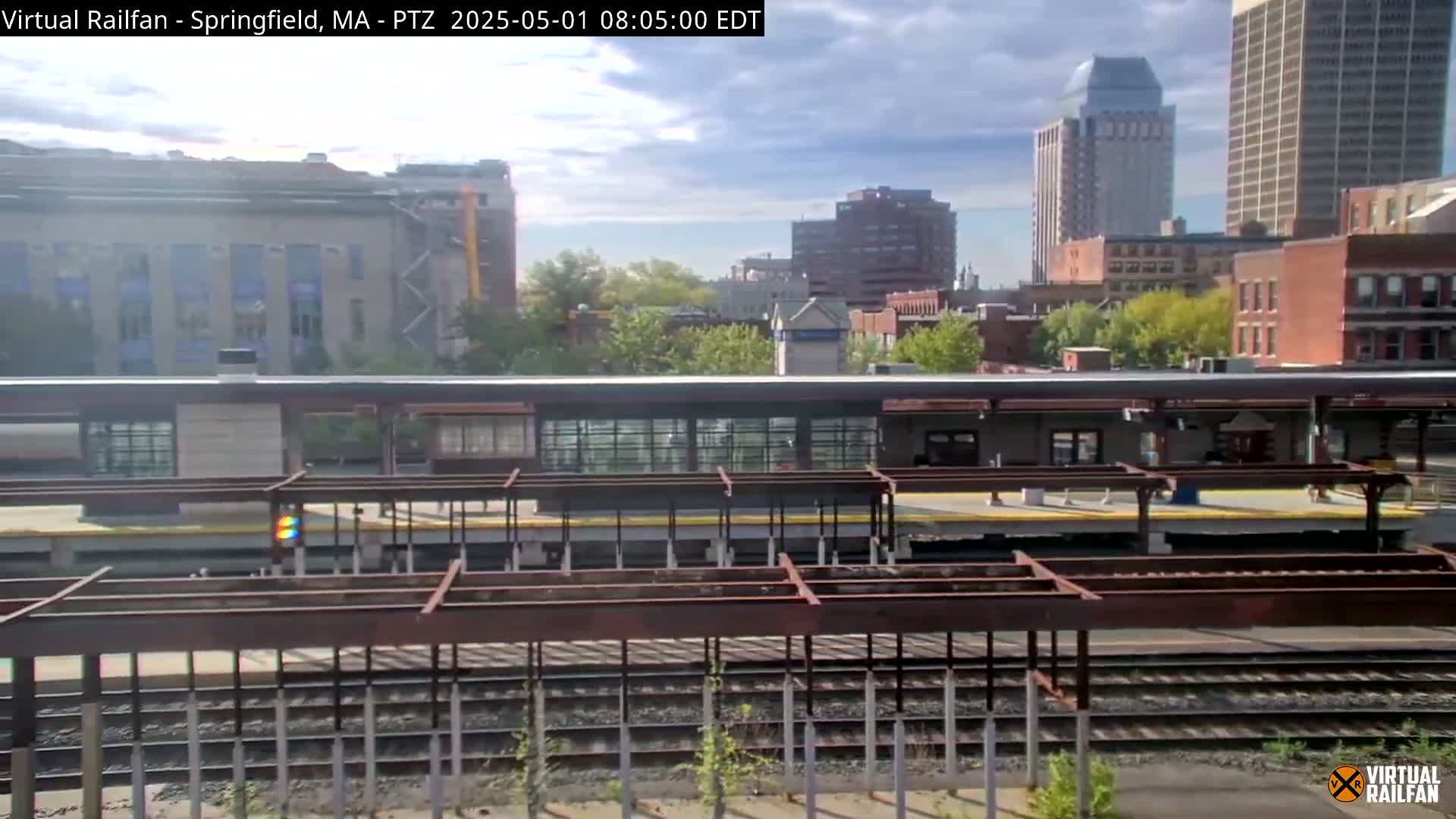 A partly sunny day shows a train station platform with several train tracks, surrounded by city buildings and trees.