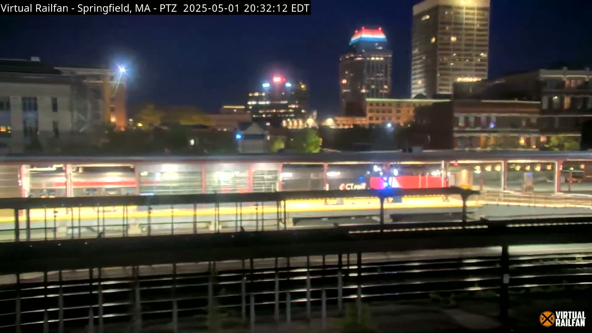 A nighttime view of a train station platform with a train at night, showing city buildings illuminated in the background.