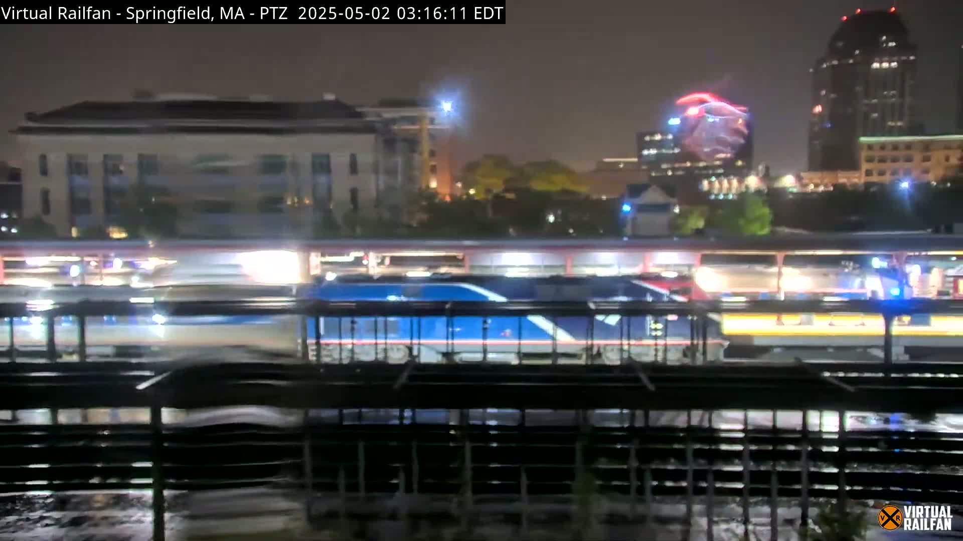 A blue and white train passes by a station at night in the rain, with city lights and buildings in the background.