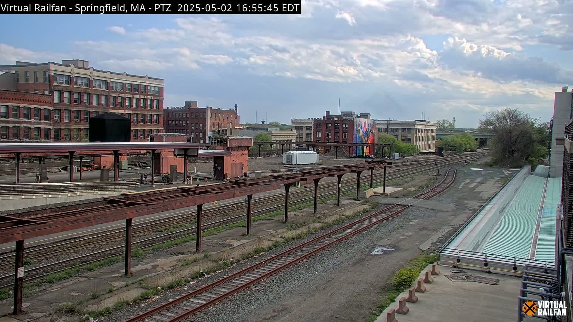 A partly cloudy day reveals a railroad yard with multiple tracks, train platforms, and brick buildings in the background.