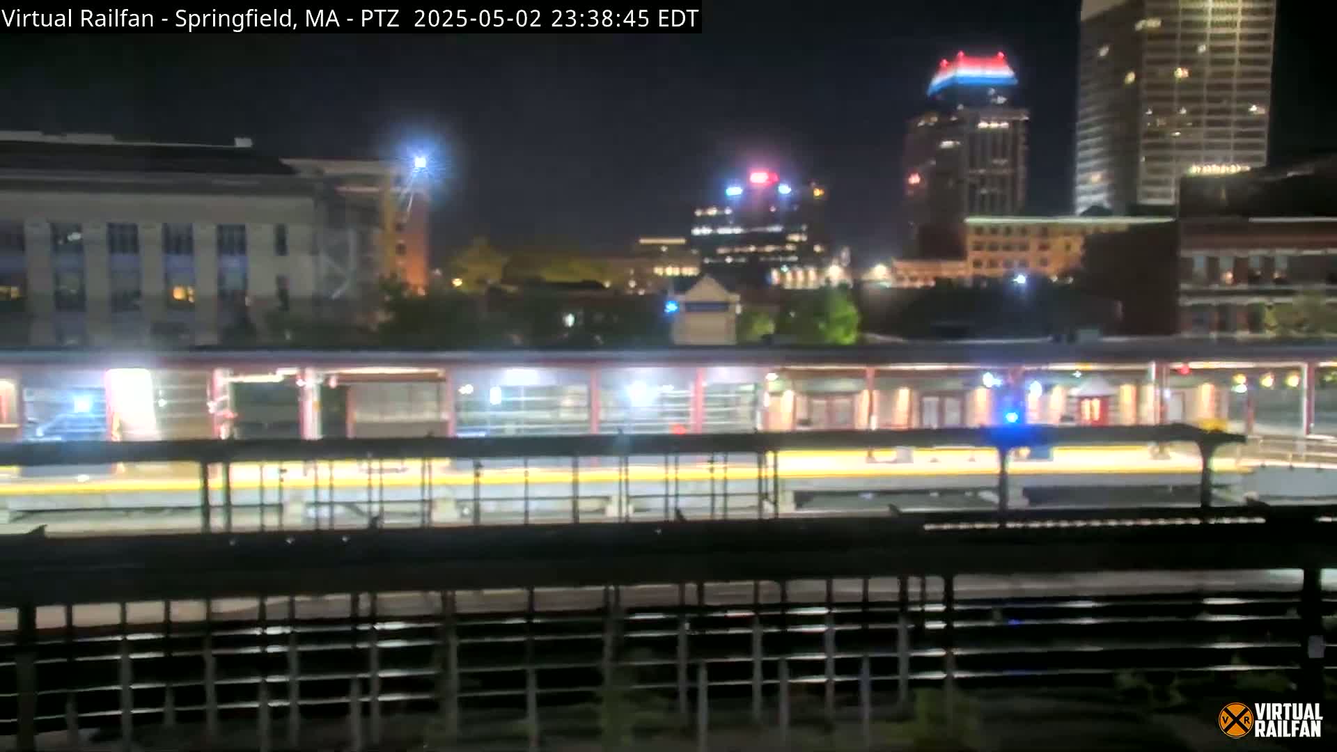 A nighttime view of a train station platform with a train at the platform, illuminated buildings in the background, and dark, clear skies.