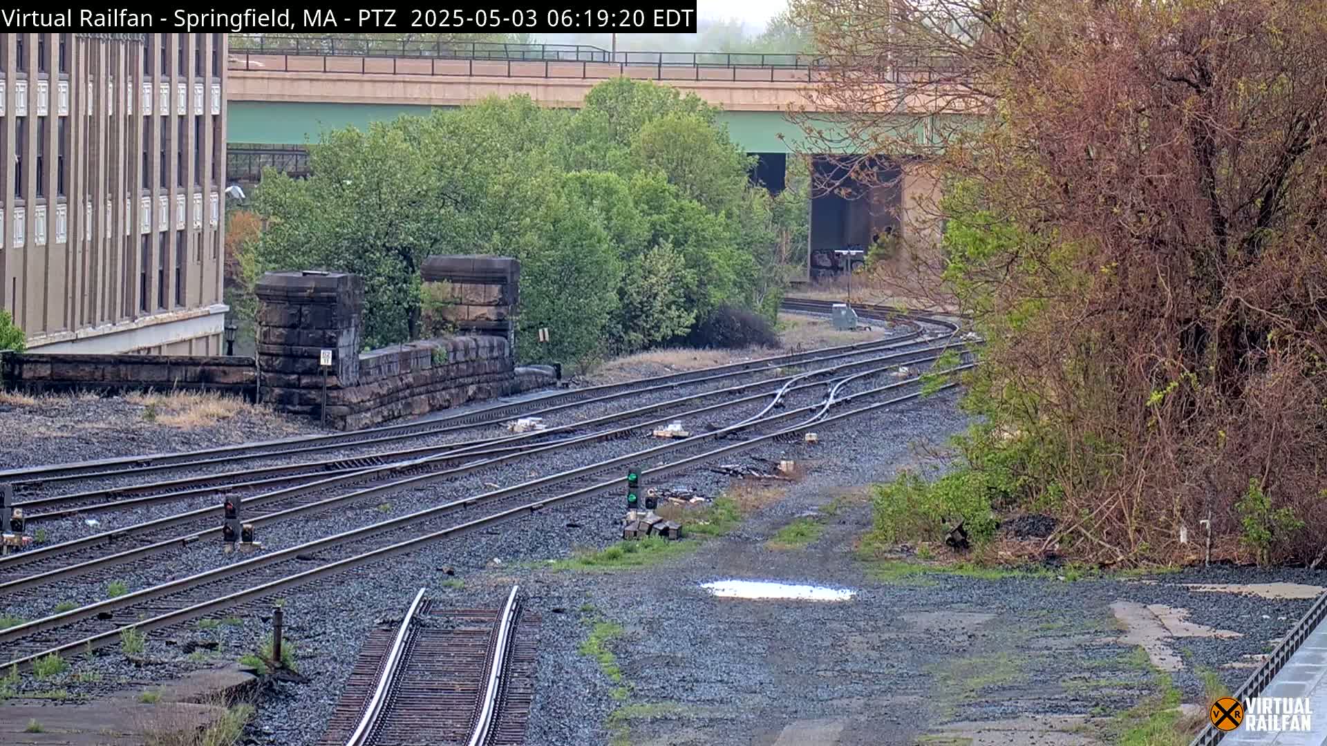 Several train tracks curve around a stone wall and under a bridge, partially obscured by vegetation in an overcast outdoor setting.