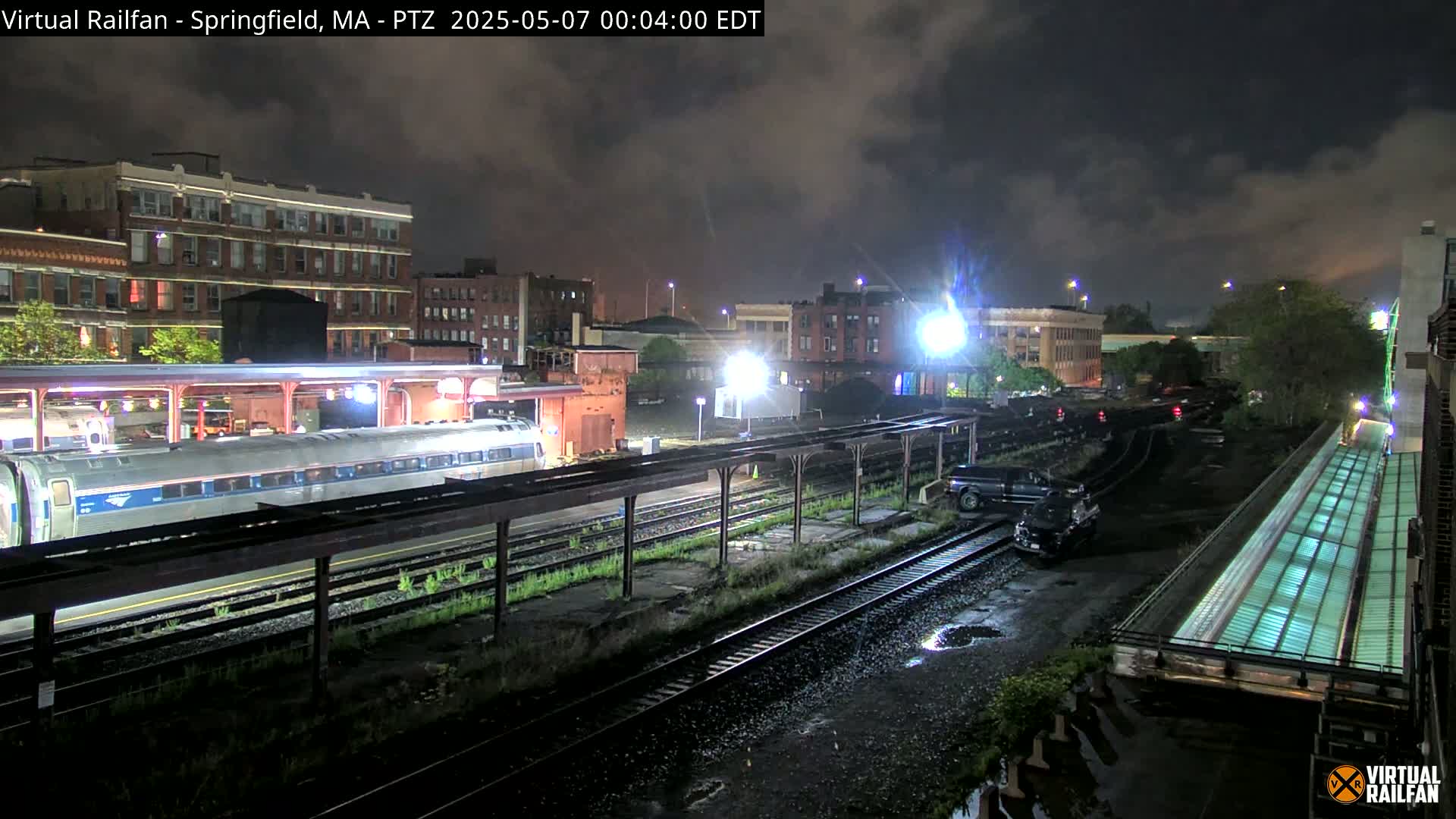 A nighttime view of a train station with a passenger train and a few vehicles, under a cloudy sky.