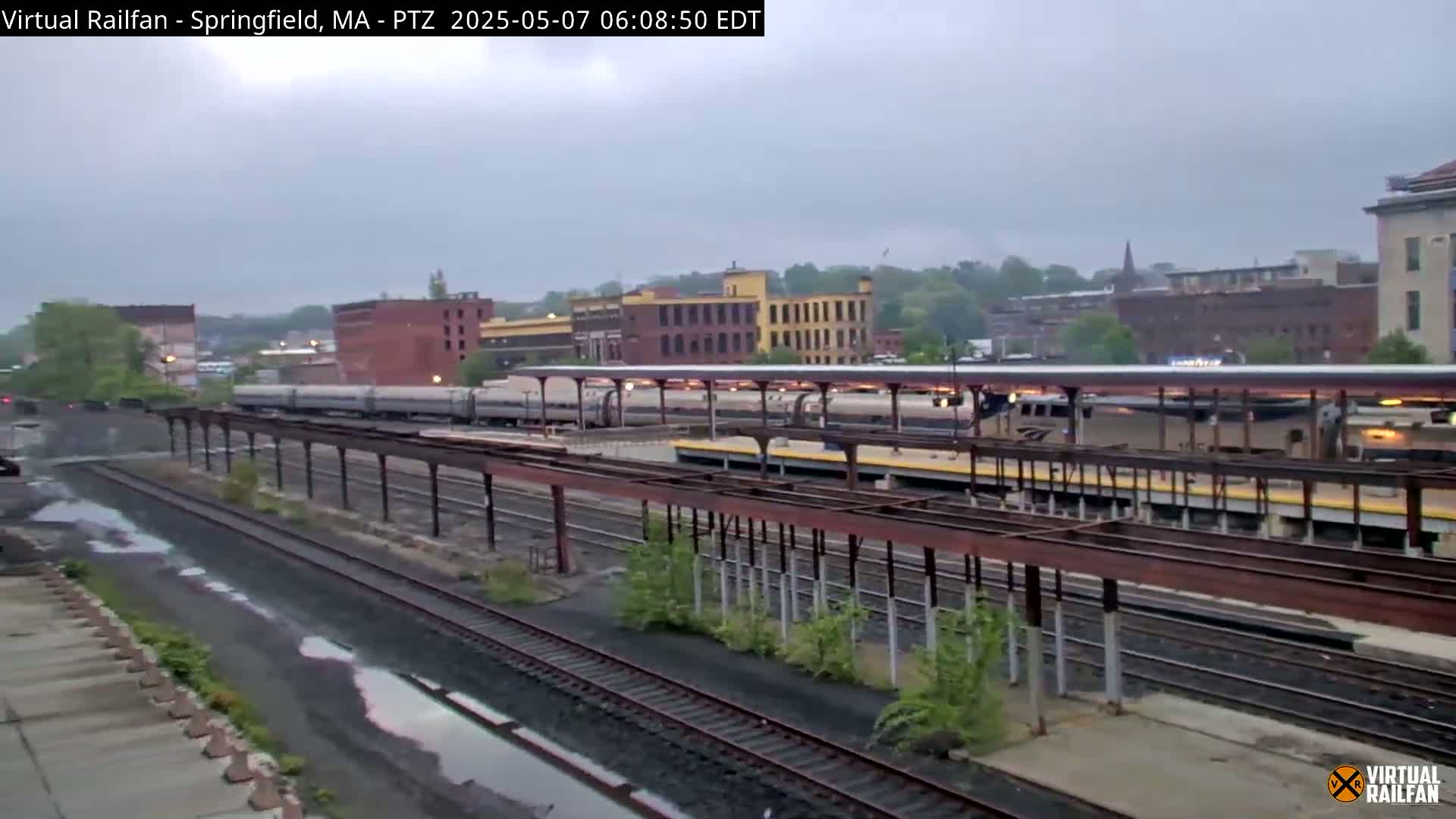 Several trains are at a station under an overcast sky, with some wetness visible on the ground.