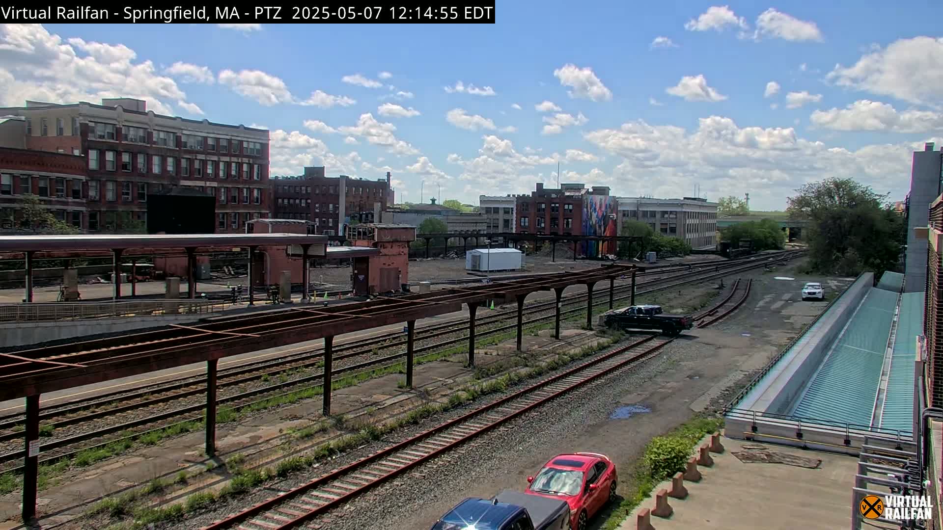 A train station with several tracks and multiple buildings in the background under a partly cloudy sky.