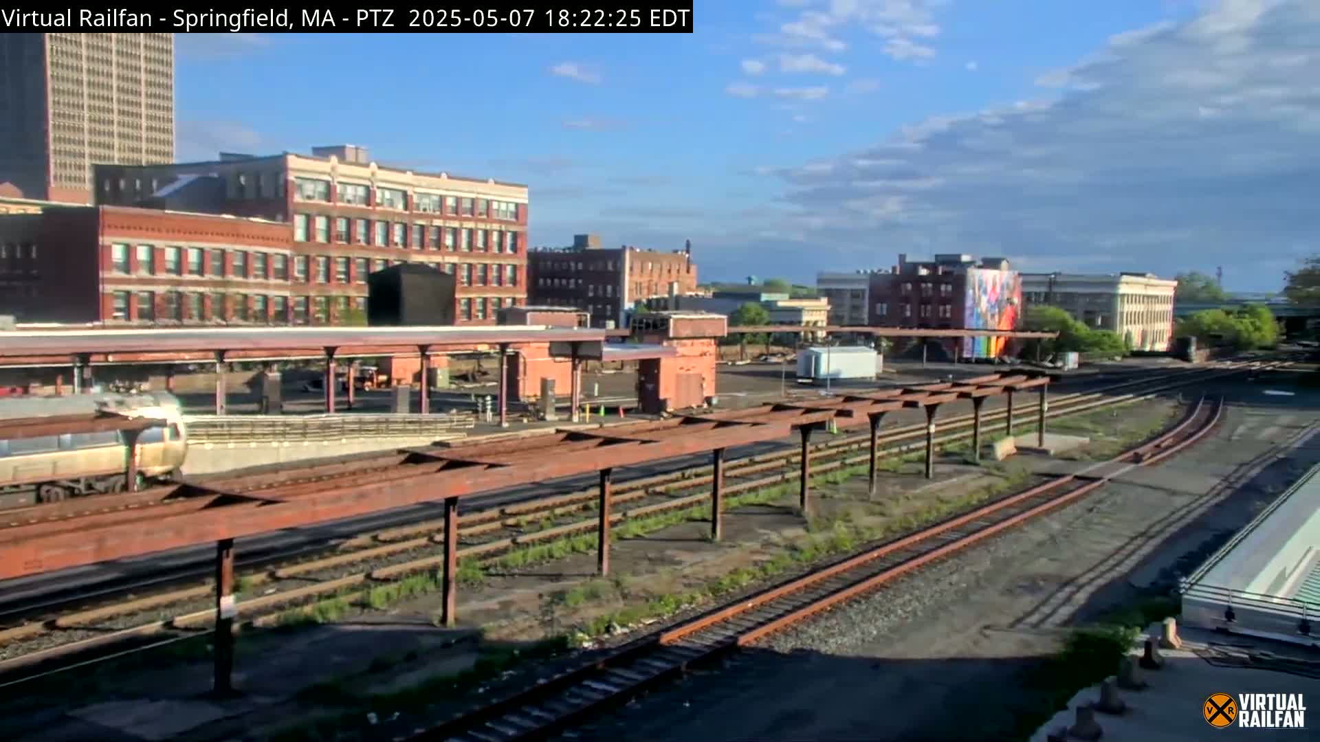 A train sits at a station next to several brick buildings under a partly cloudy sky.