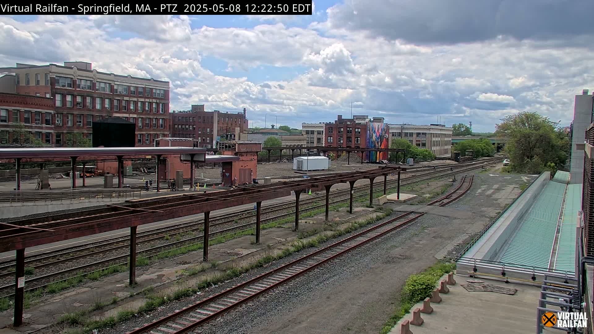 A mostly cloudy day reveals a train yard with several tracks, a partially-demolished building, and brick buildings in the background.